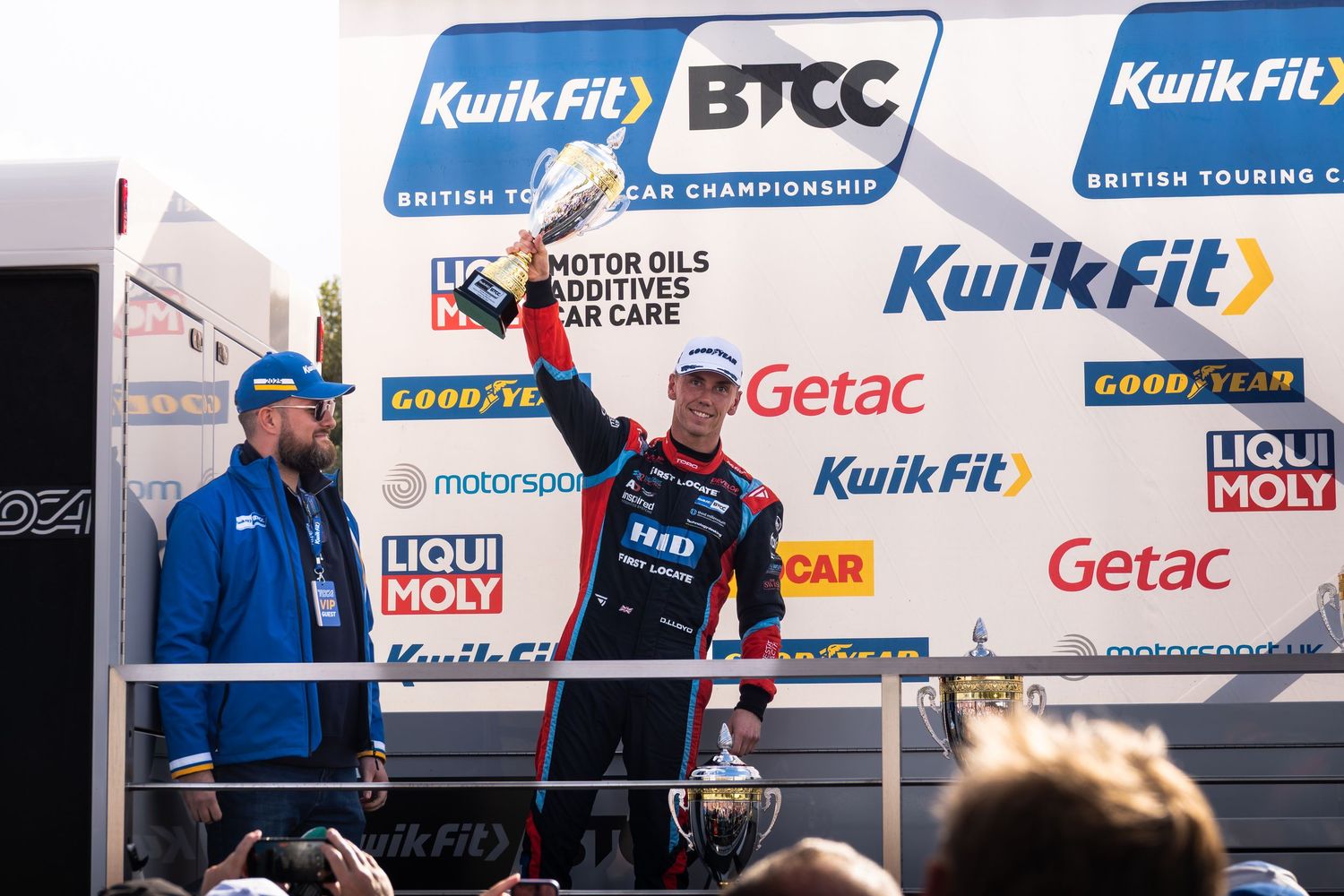 A racing driver celebrates on the KwikFit BTCC British Touring Car Championship podium holding a trophy.