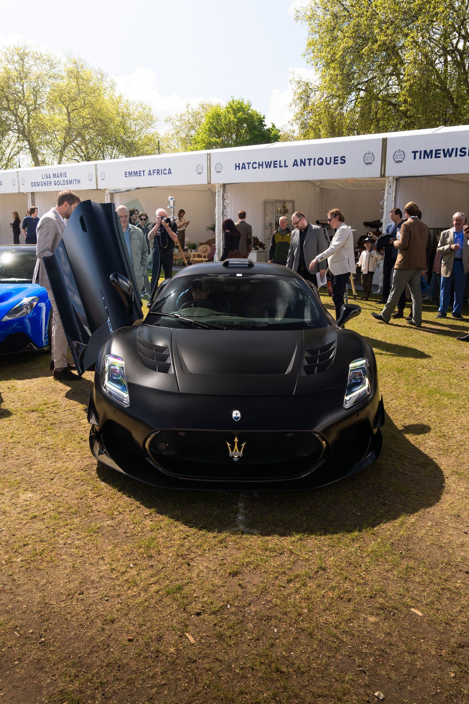 Matte black Maserati MC20 supercar with scissor doors open on display at an outdoor luxury car show.