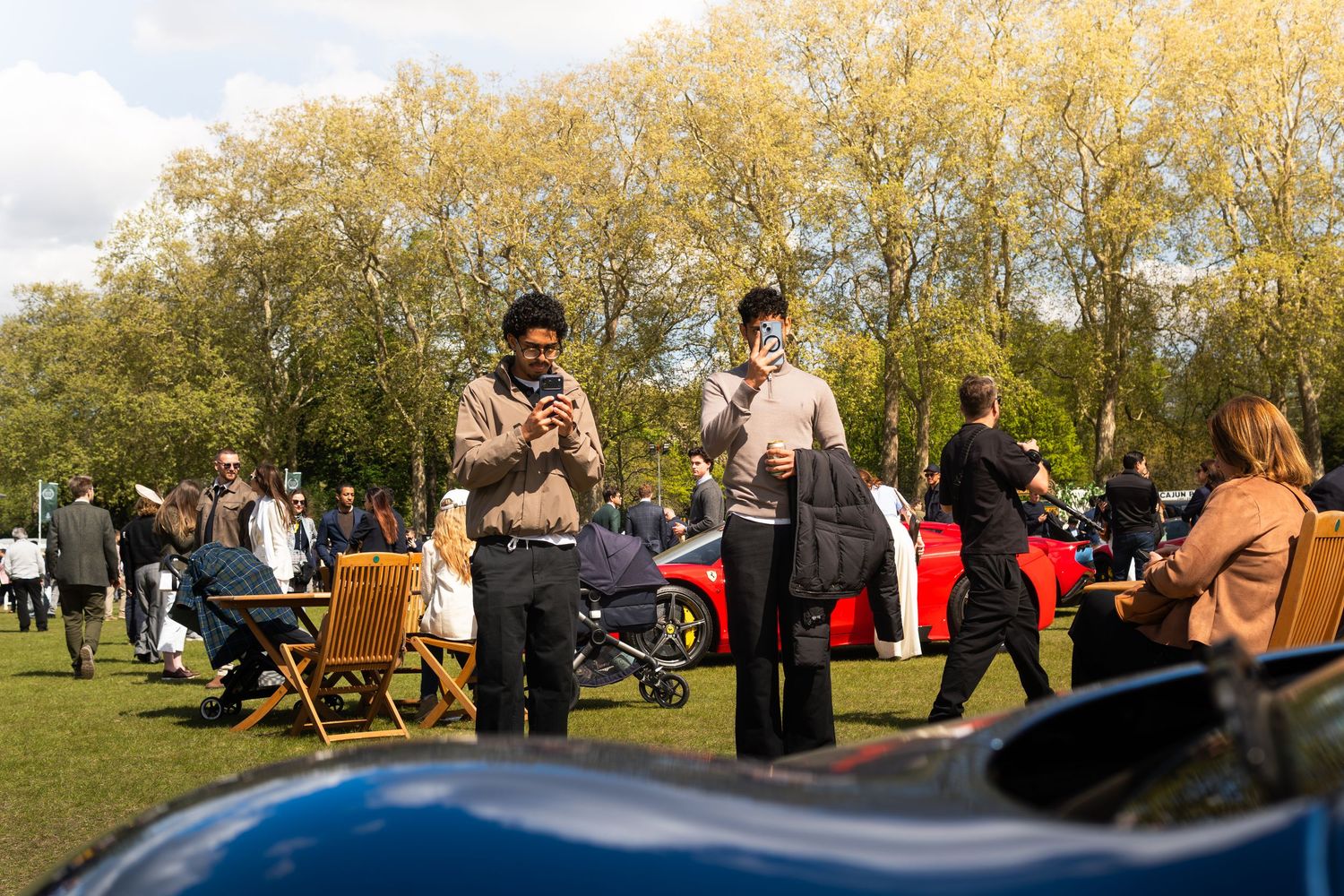 Visitors using smartphones at an outdoor exotic car show surrounded by trees and parked supercars.