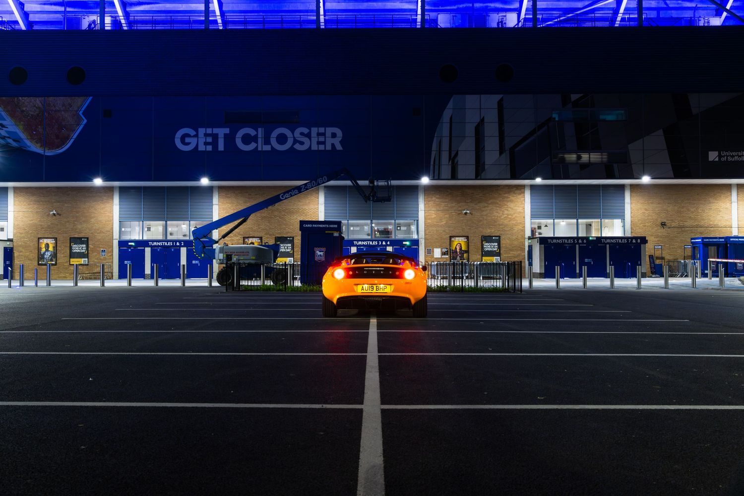 Orange Lotus Elise parked at night outside Ipswich Town Football Club stadium with blue illuminated GET CLOSER signage.