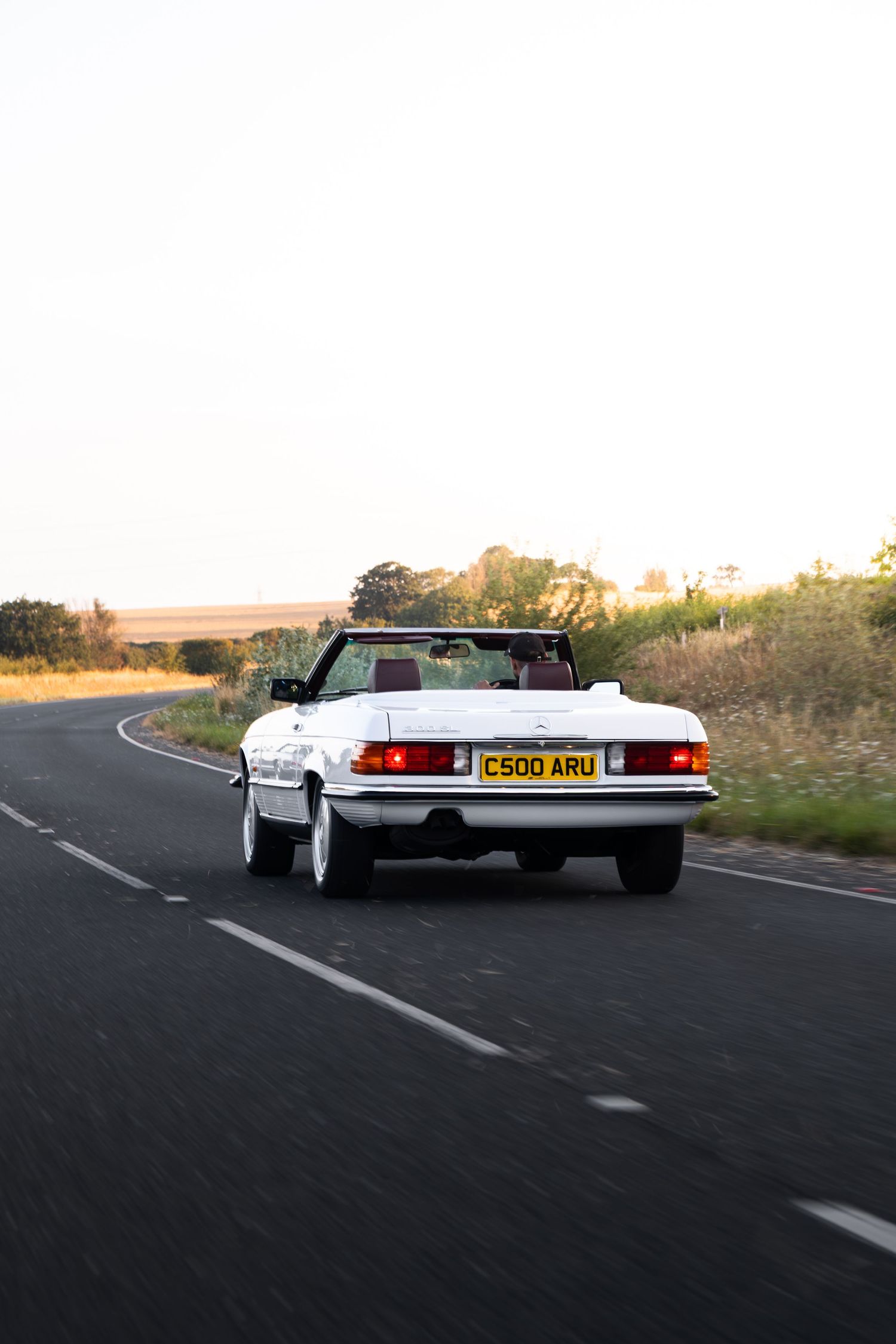 White convertible Mercedes-Benz SL driving on a rural road, rear view showing UK license plate C500 ARU at dusk.