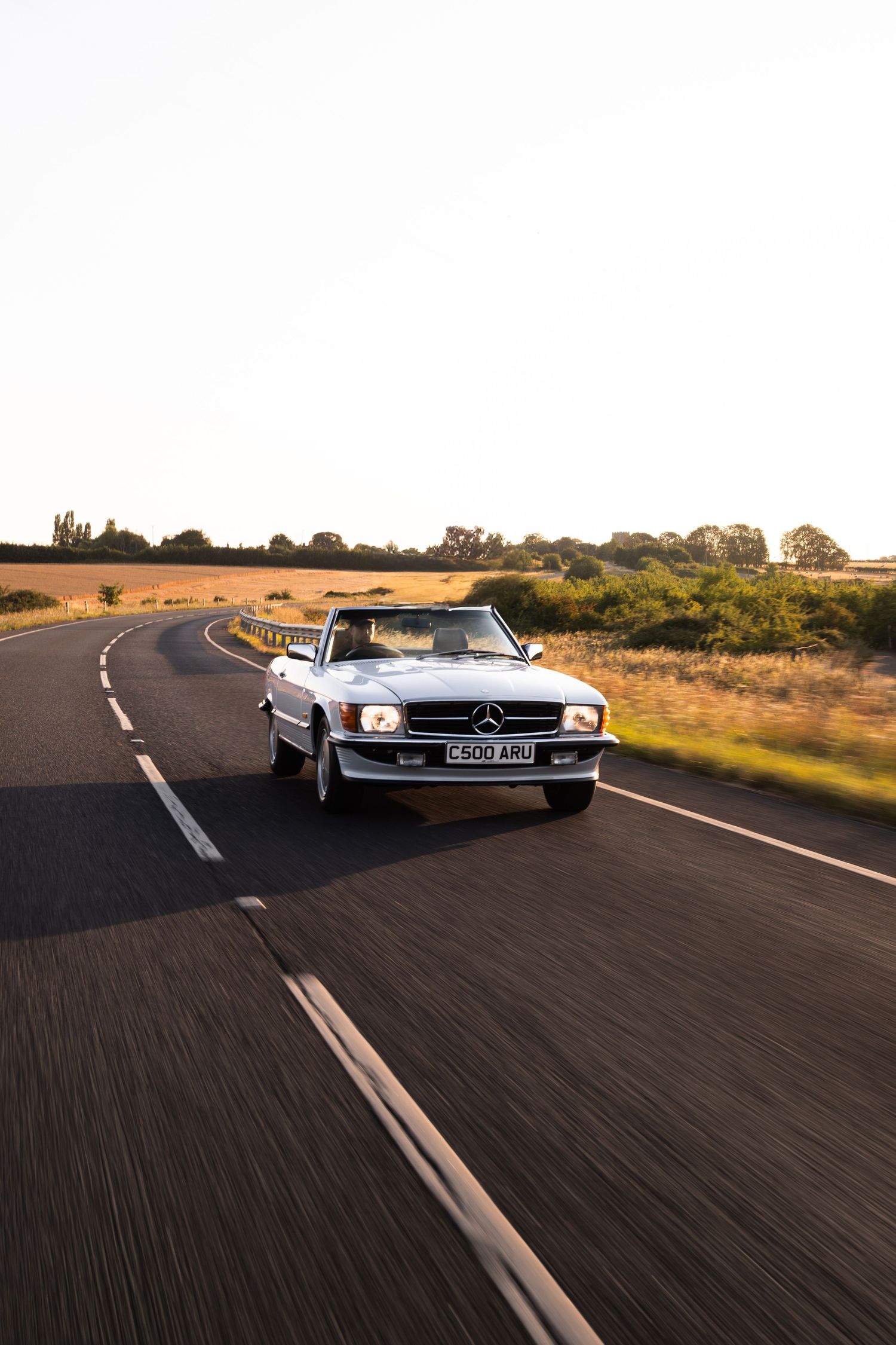 A classic Mercedes-Benz convertible drives along a scenic rural road at golden hour, surrounded by fields.