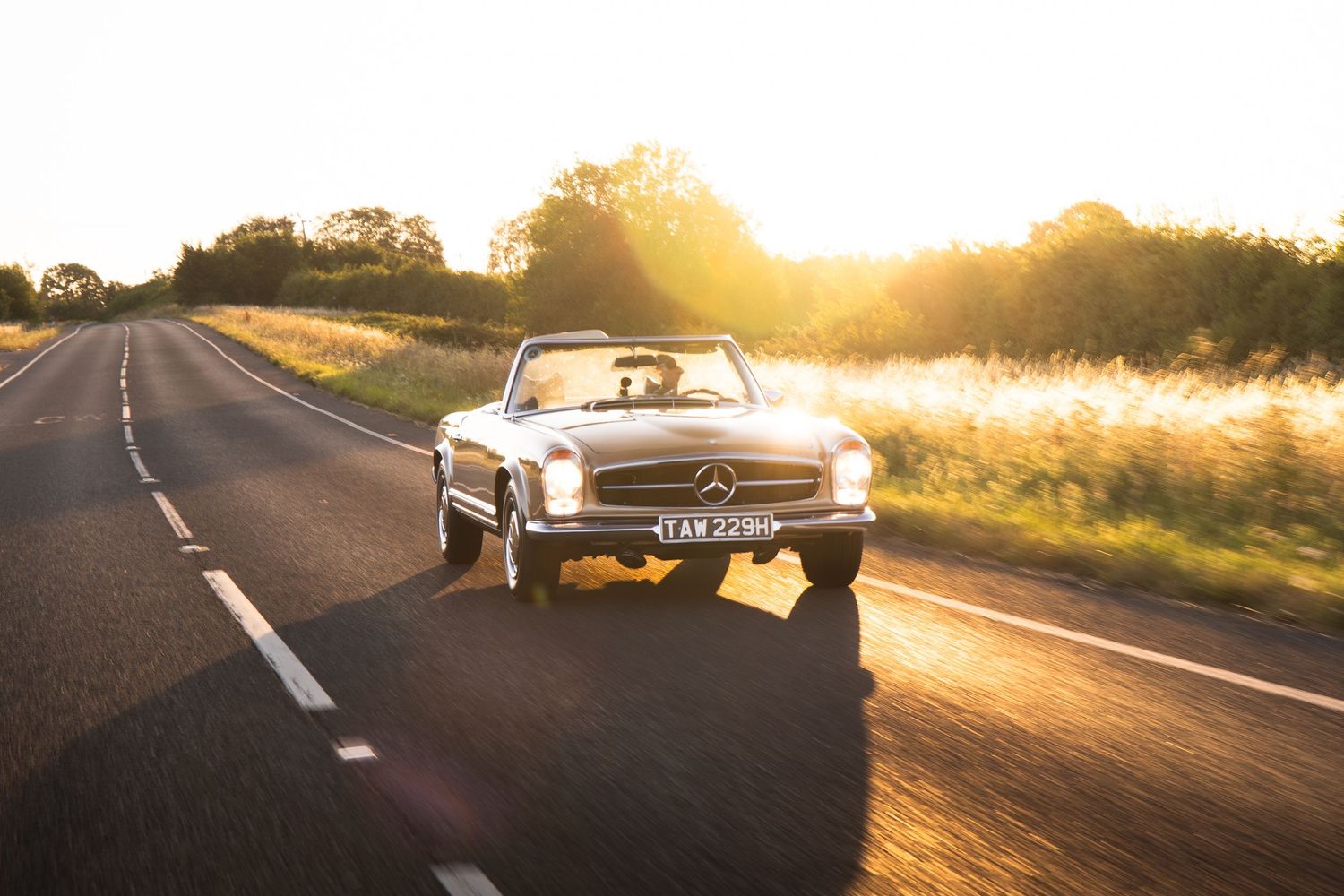 Classic Mercedes-Benz convertible driving on a scenic road at golden sunset, surrounded by lush green fields.