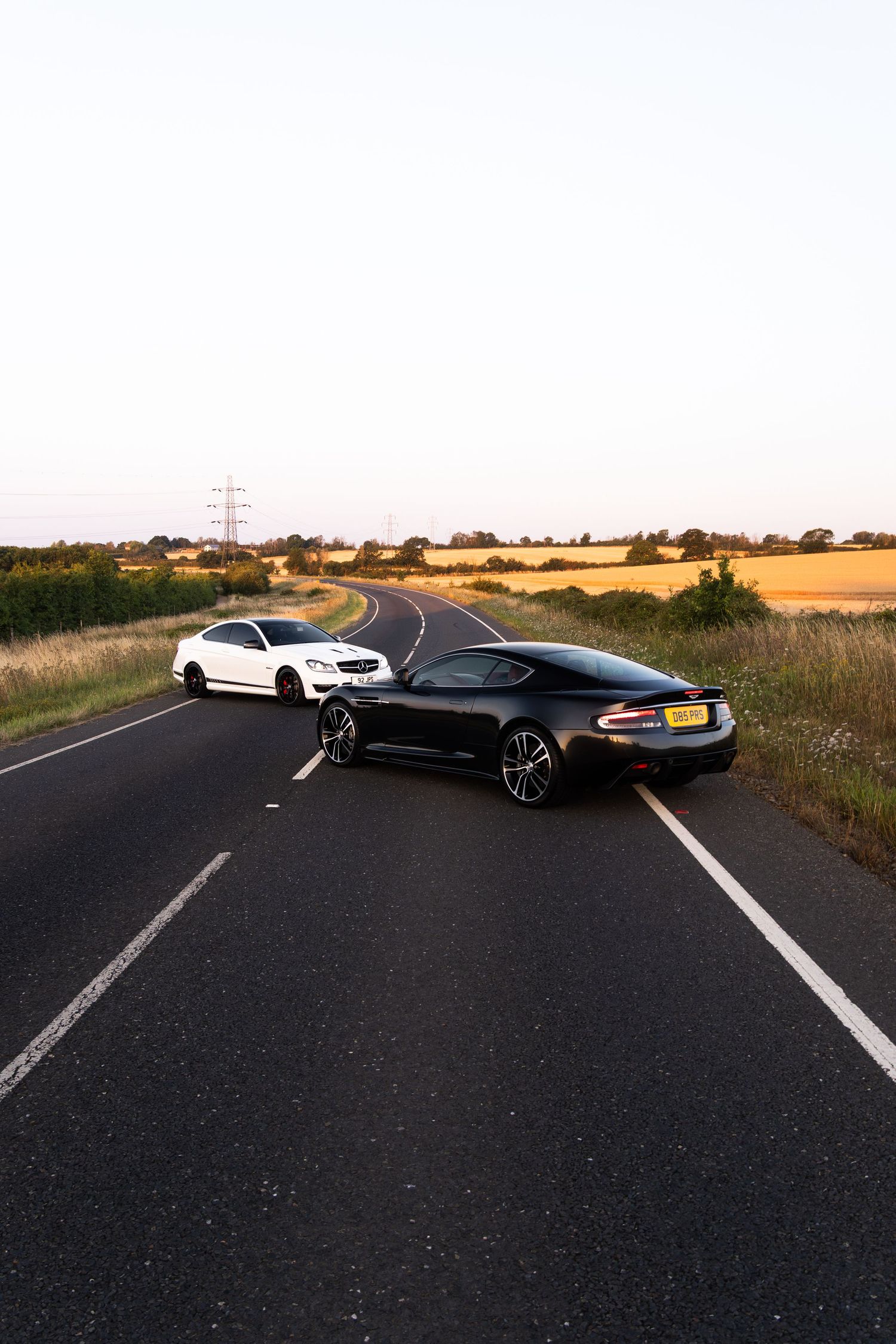 Black Aston Martin DBS and white Mercedes coupe parked on rural English road at golden hour, surrounded by fields.