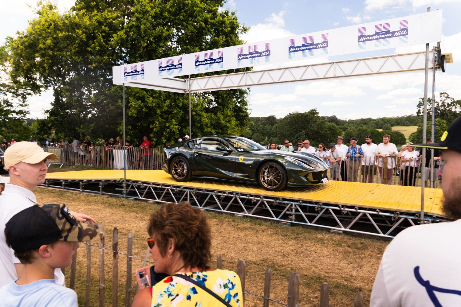 Dark green supercar displayed on a yellow platform at Goodwood Festival of Speed, surrounded by spectators under a sunny sky.