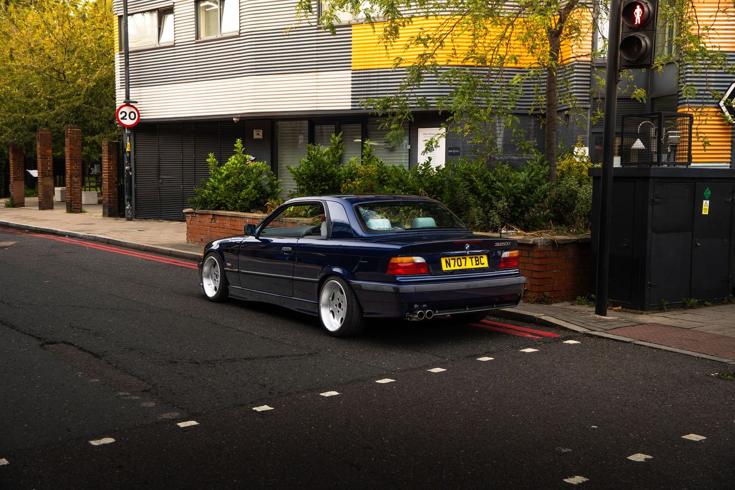 A lowered blue BMW E36 coupe on silver wheels stopped at a traffic light on a London street.