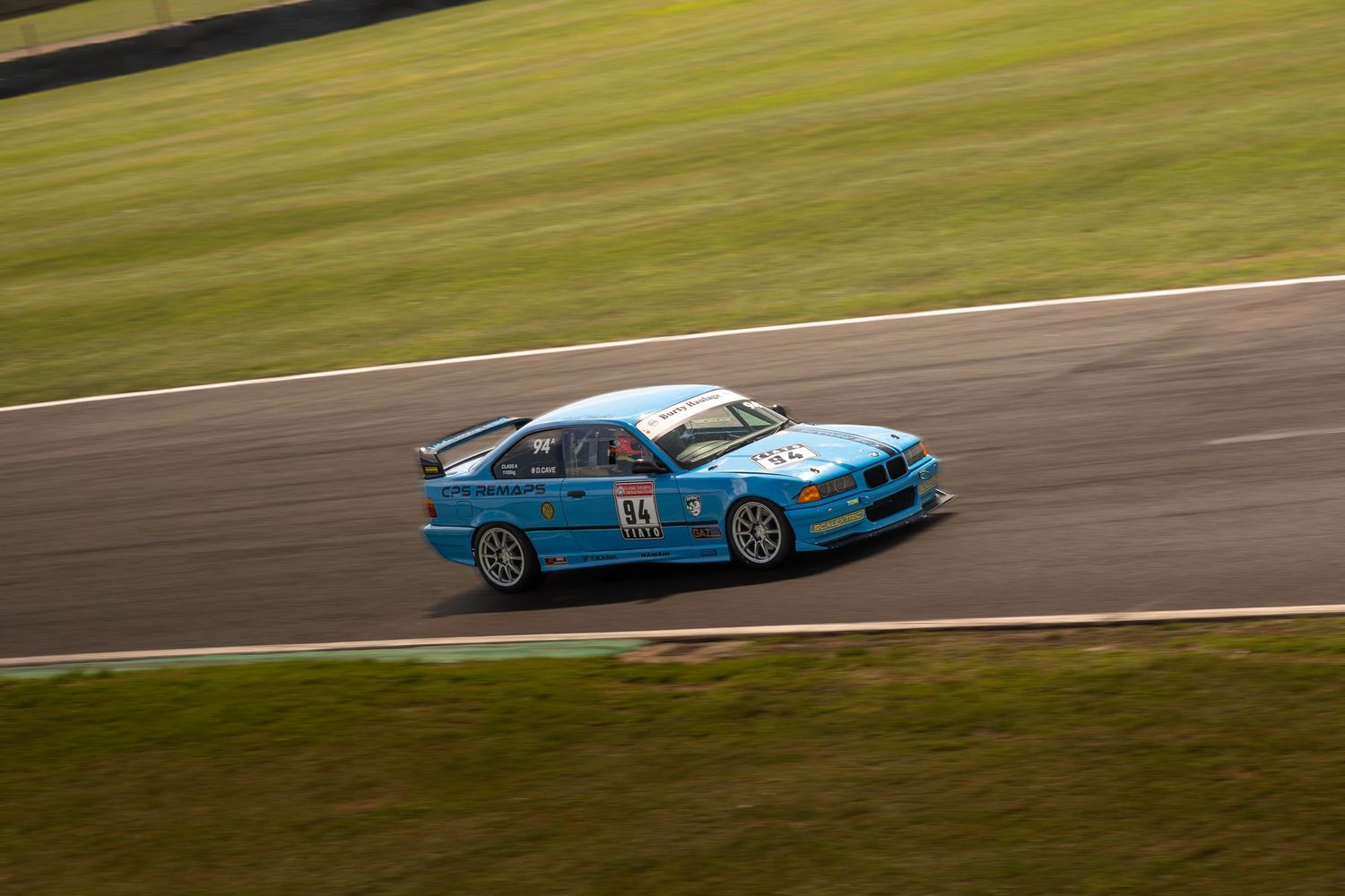 Blue BMW touring car number 64 racing on a sunlit motorsport circuit.