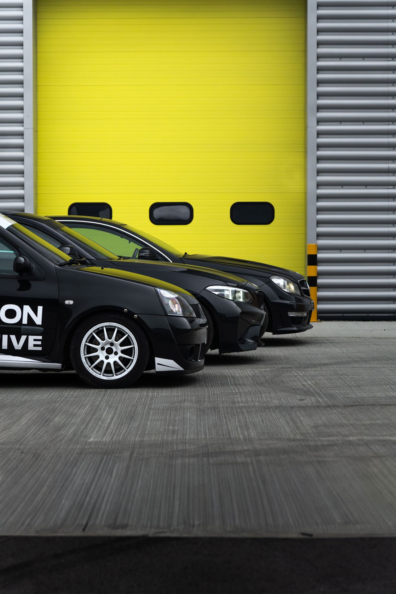 Two black Motion Automotive performance cars parked in front of a bright yellow industrial garage door.
