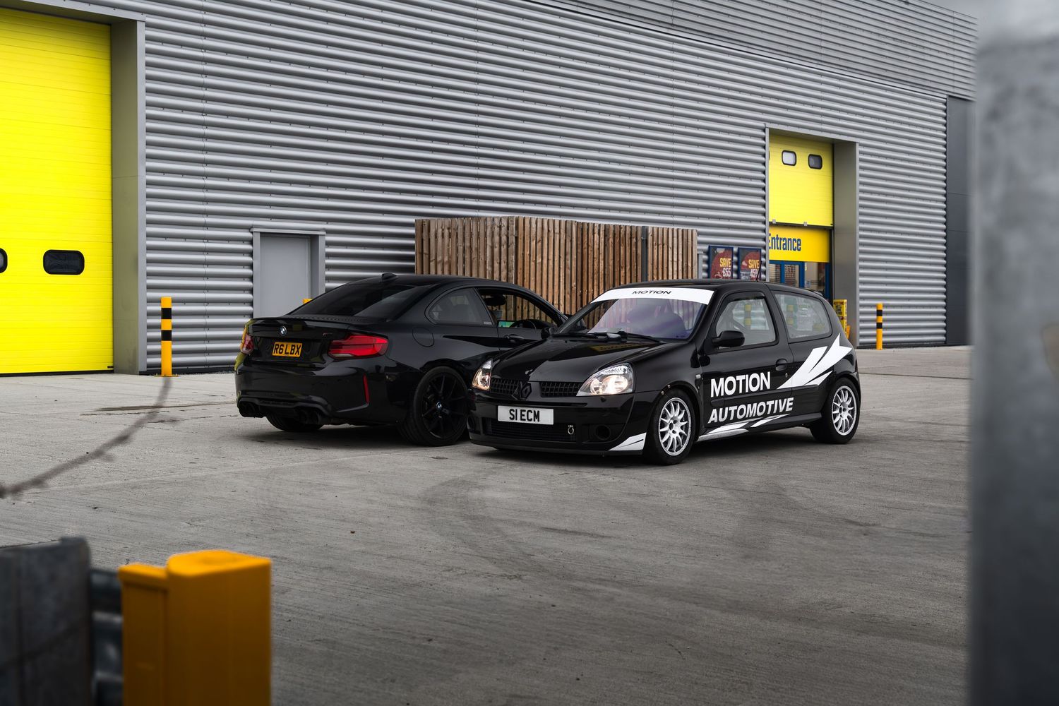 Black BMW and Motion Automotive branded VW Golf parked outside industrial garage.