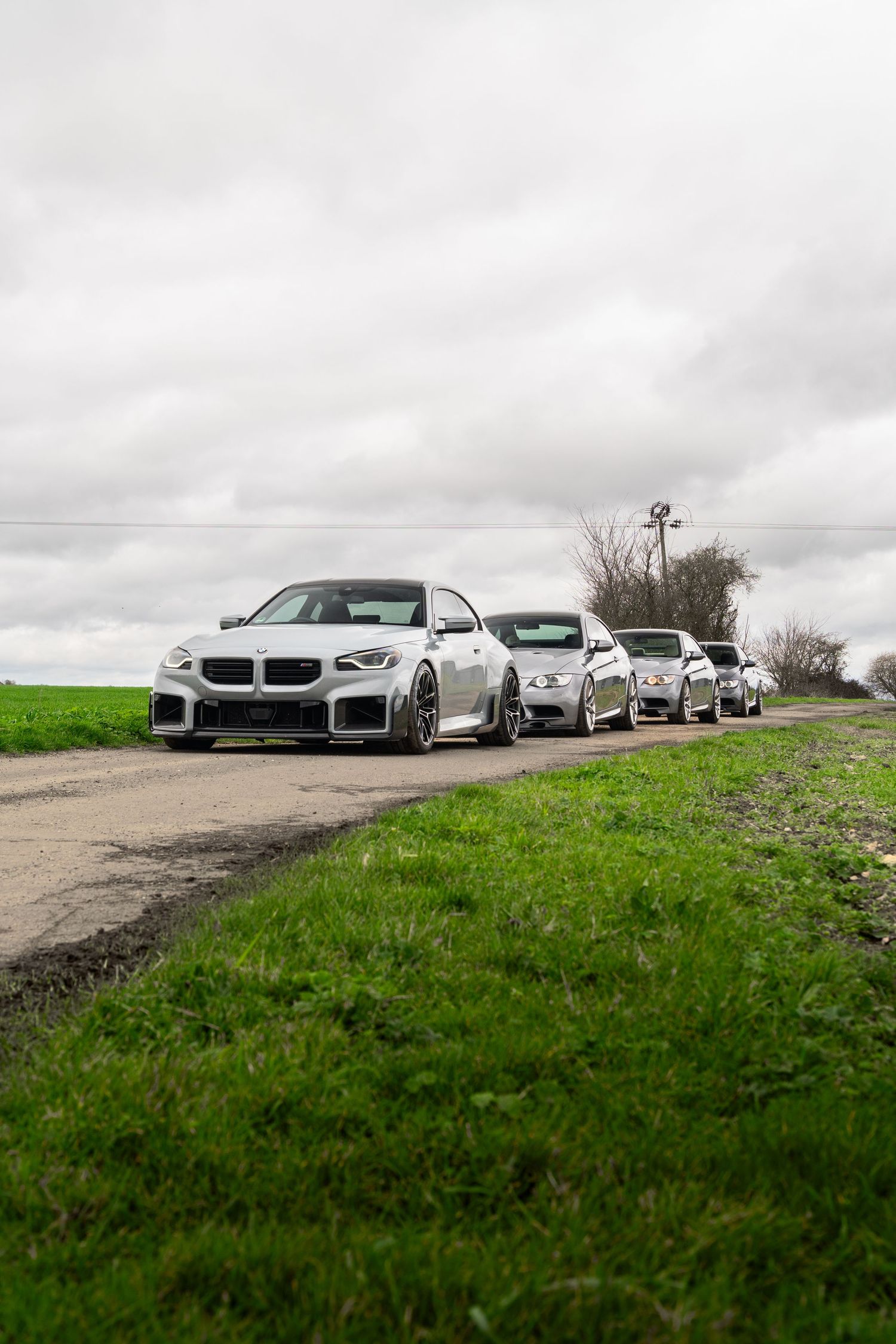 Line of silver BMW performance coupes on a rural road flanked by green fields.