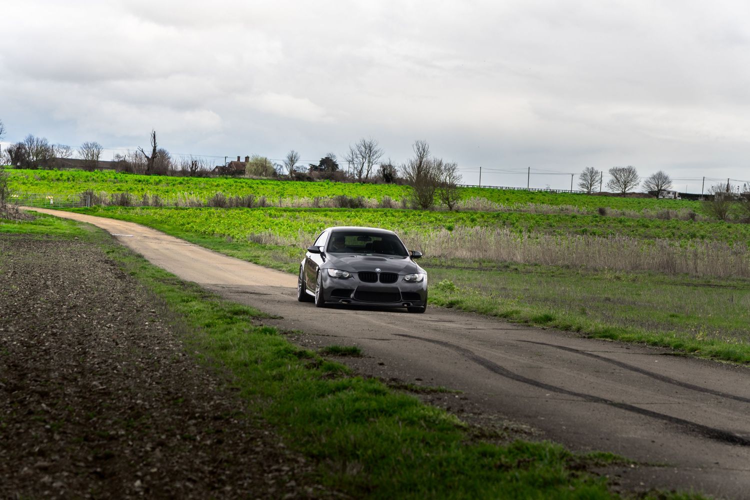 Silver BMW M3 coupe driving along a narrow rural road surrounded by green fields under an overcast sky.