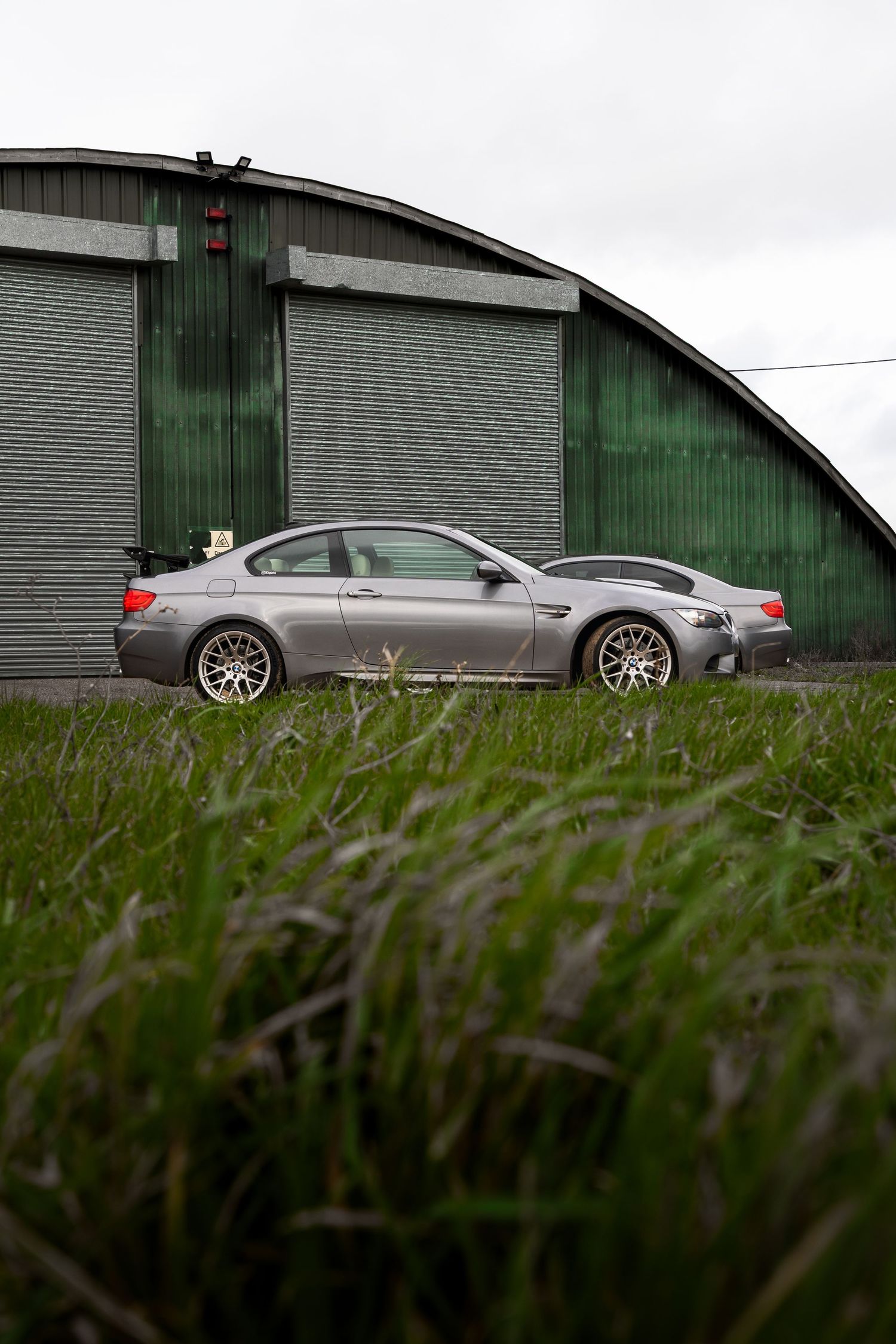 Side profile of a gray BMW M3 E92 coupe parked beside a curved green hangar, shot through grass.