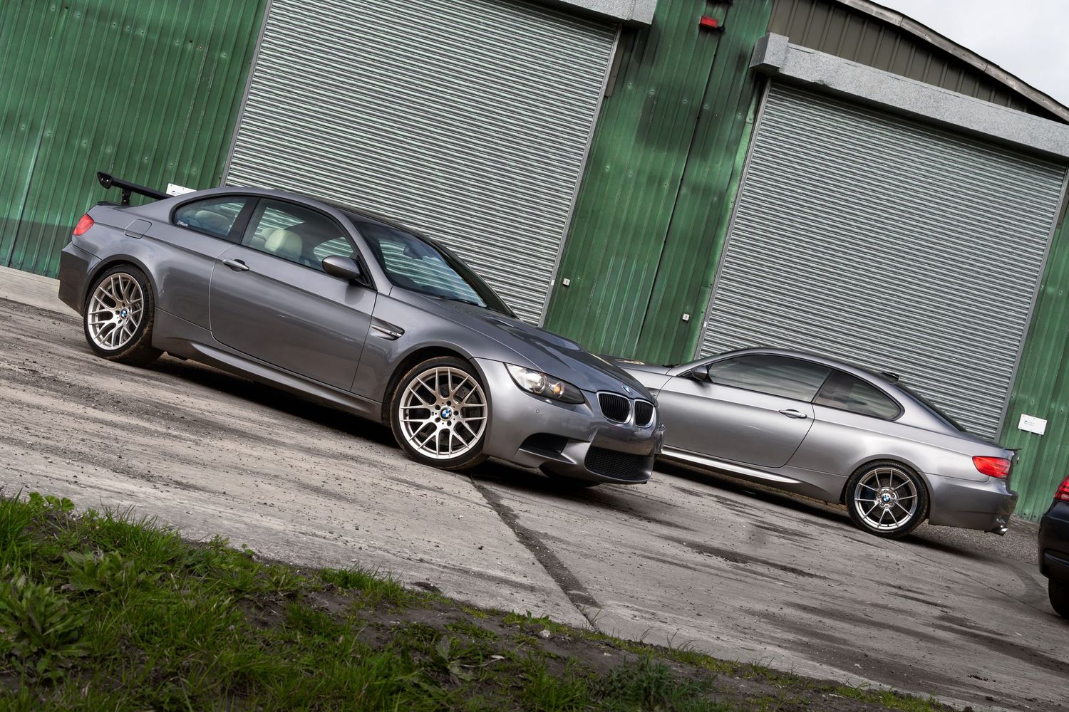 Two gray BMW M3 coupes parked side by side in front of green industrial garage doors.