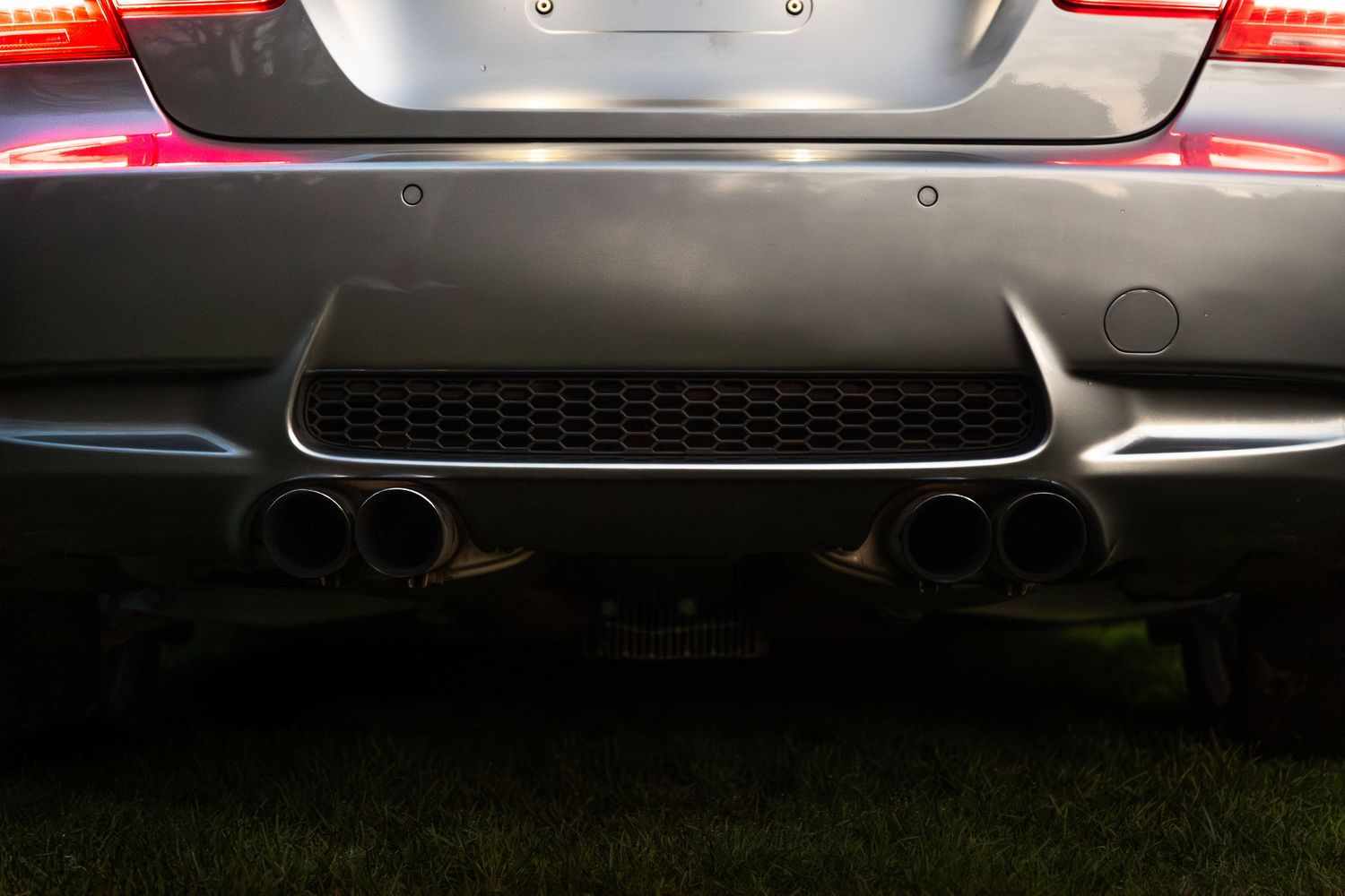 Rear diffuser and quad exhaust tips of a grey BMW M3 illuminated by tail lights on grass.