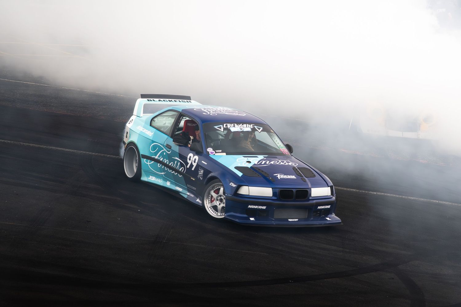 A blue BMW drift car enveloped in thick white tire smoke performs a drift on a dark racing track.