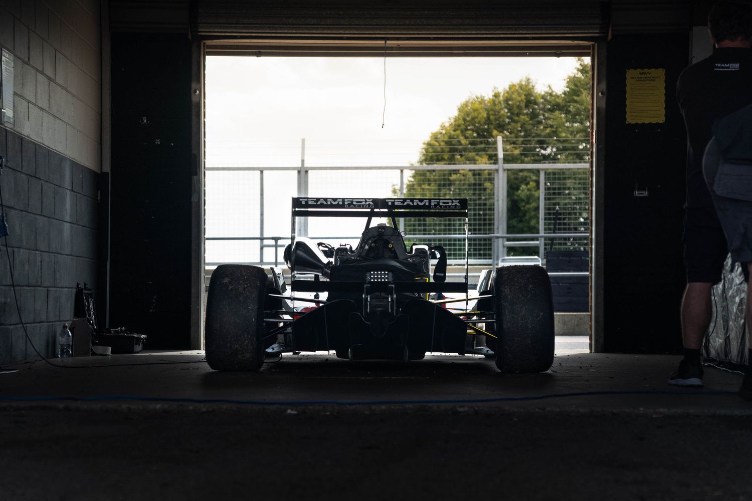 A dark silhouette of a Formula racing car sits inside a dimly lit garage at a racing circuit.