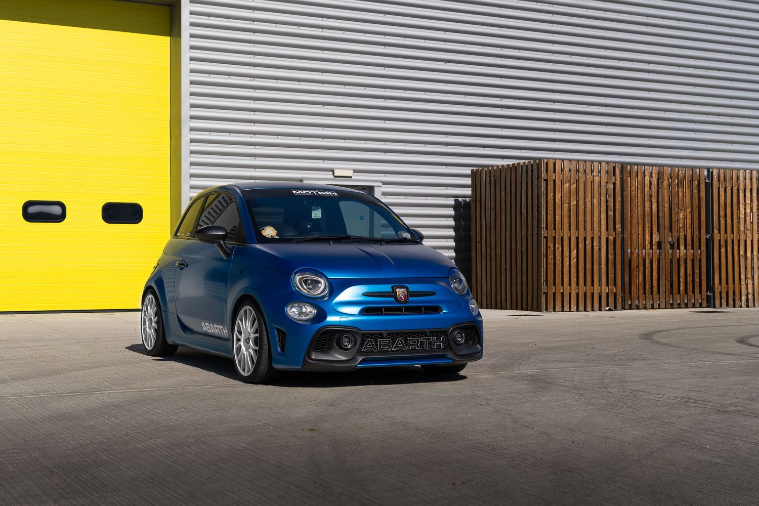 Blue Abarth 500 modified hatchback parked in front of yellow industrial garage doors.