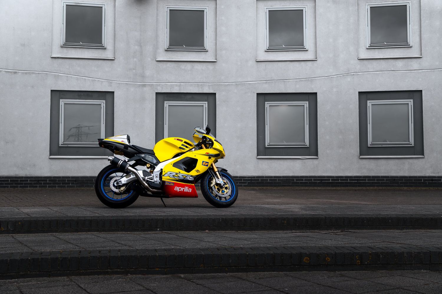 Yellow Aprilia RS5 sport motorcycle parked in front of a grey building with symmetrical windows.