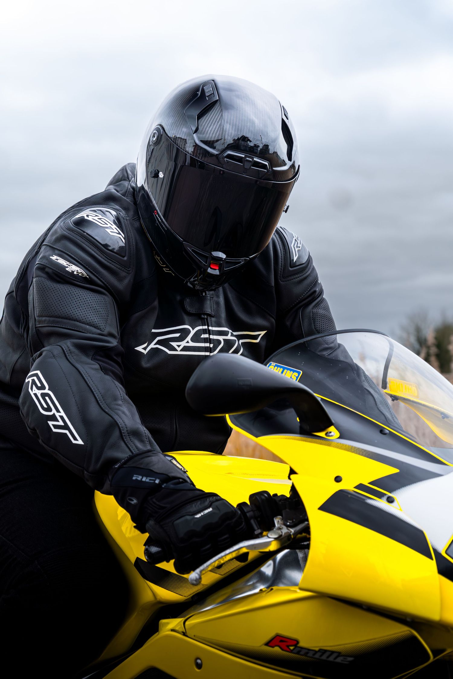 Motorcyclist in RST black leather jacket and helmet sitting on a yellow Aprilia sport bike.