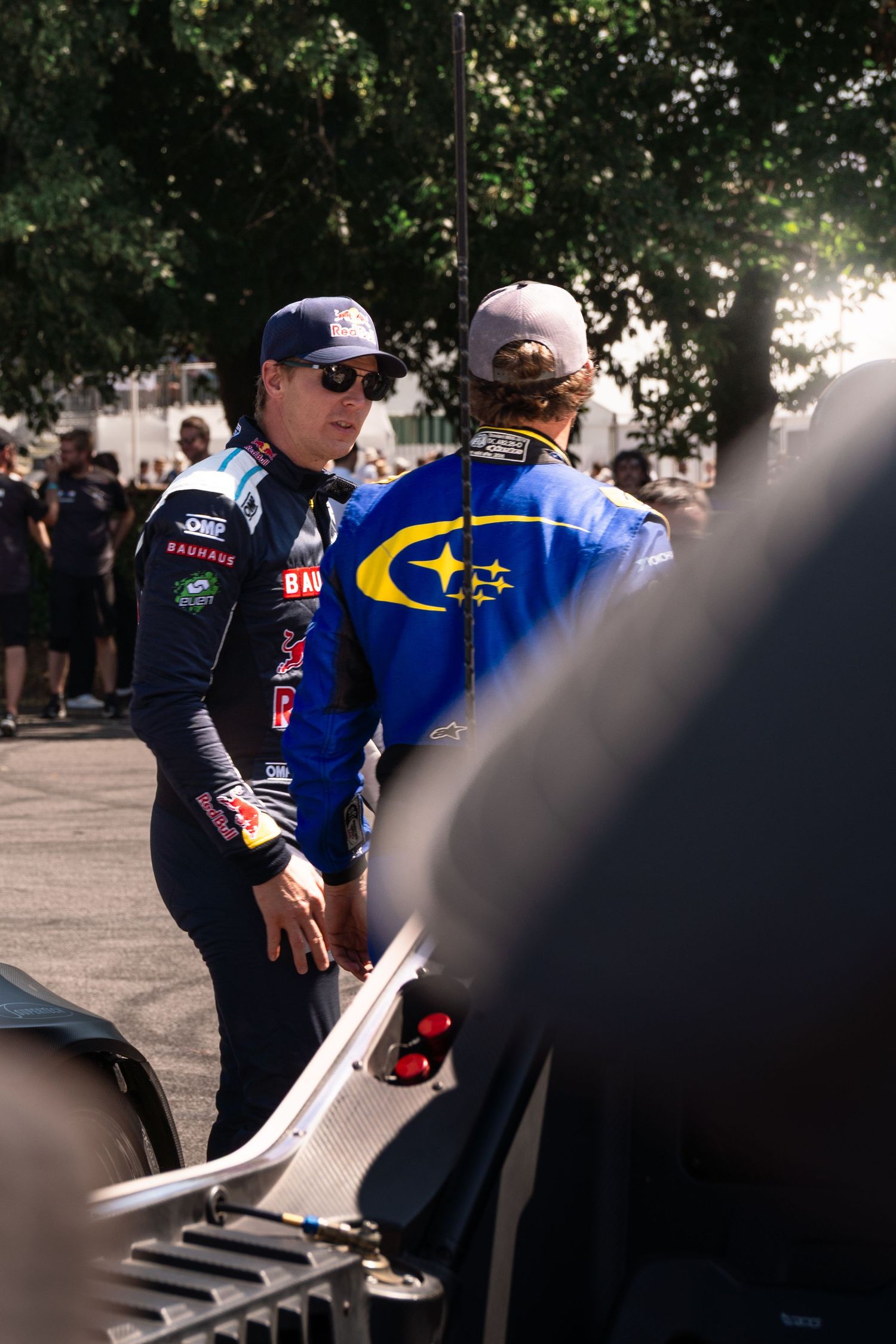 Two racing drivers in branded suits chat at an outdoor motorsport event, one wearing Subaru livery.