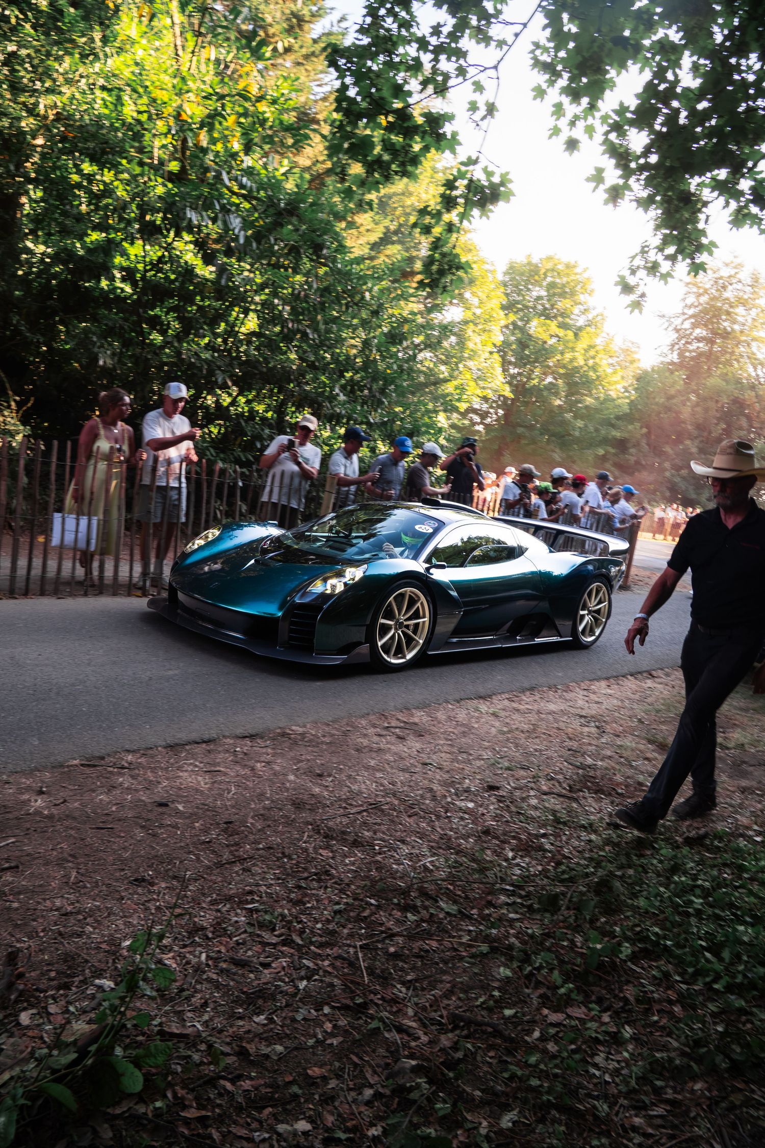 A sleek green Pagani supercar drives past spectators lining a tree-lined road at an outdoor motorsport event.