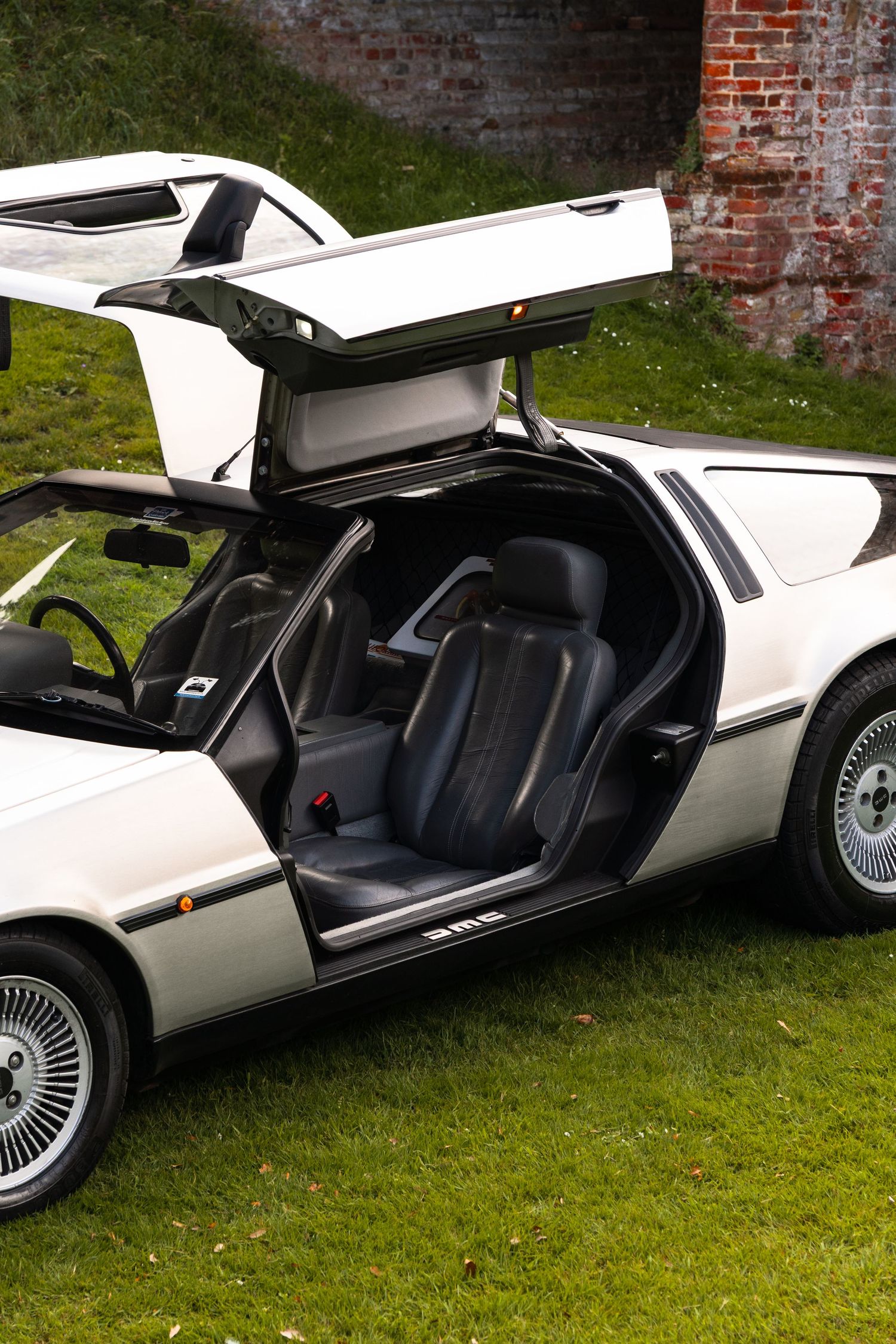 DeLorean DMC-12 with gullwing doors open, showing black interior, parked on grass near brick wall.