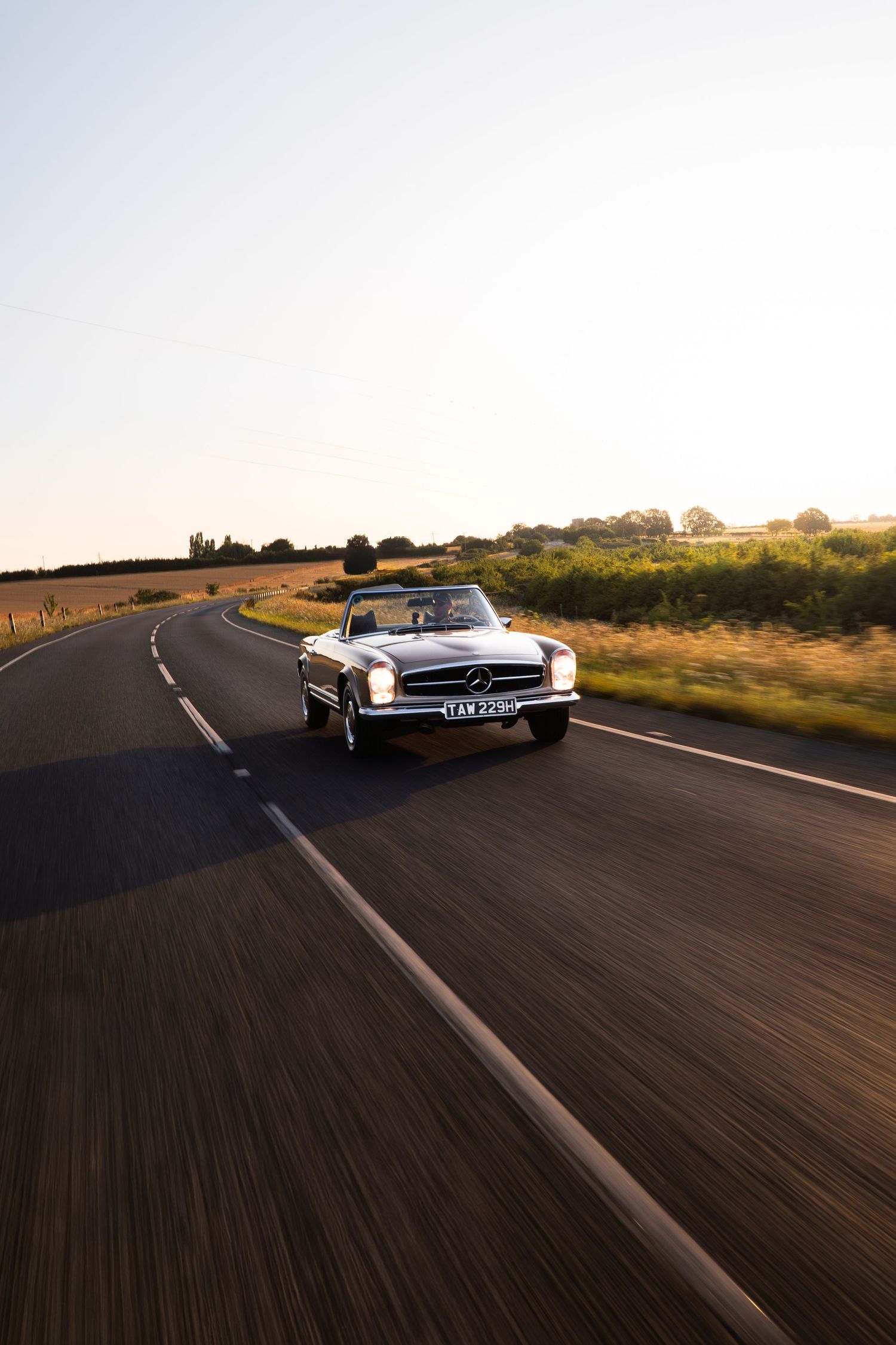 Classic Mercedes-Benz convertible driving on a scenic rural road at golden hour with motion blur effect.