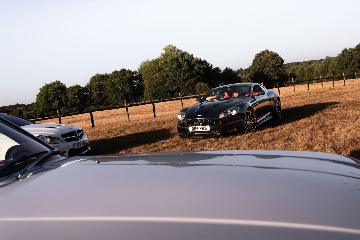 Two luxury sports cars parked on a dry grass field near wooden fencing with trees in the background.