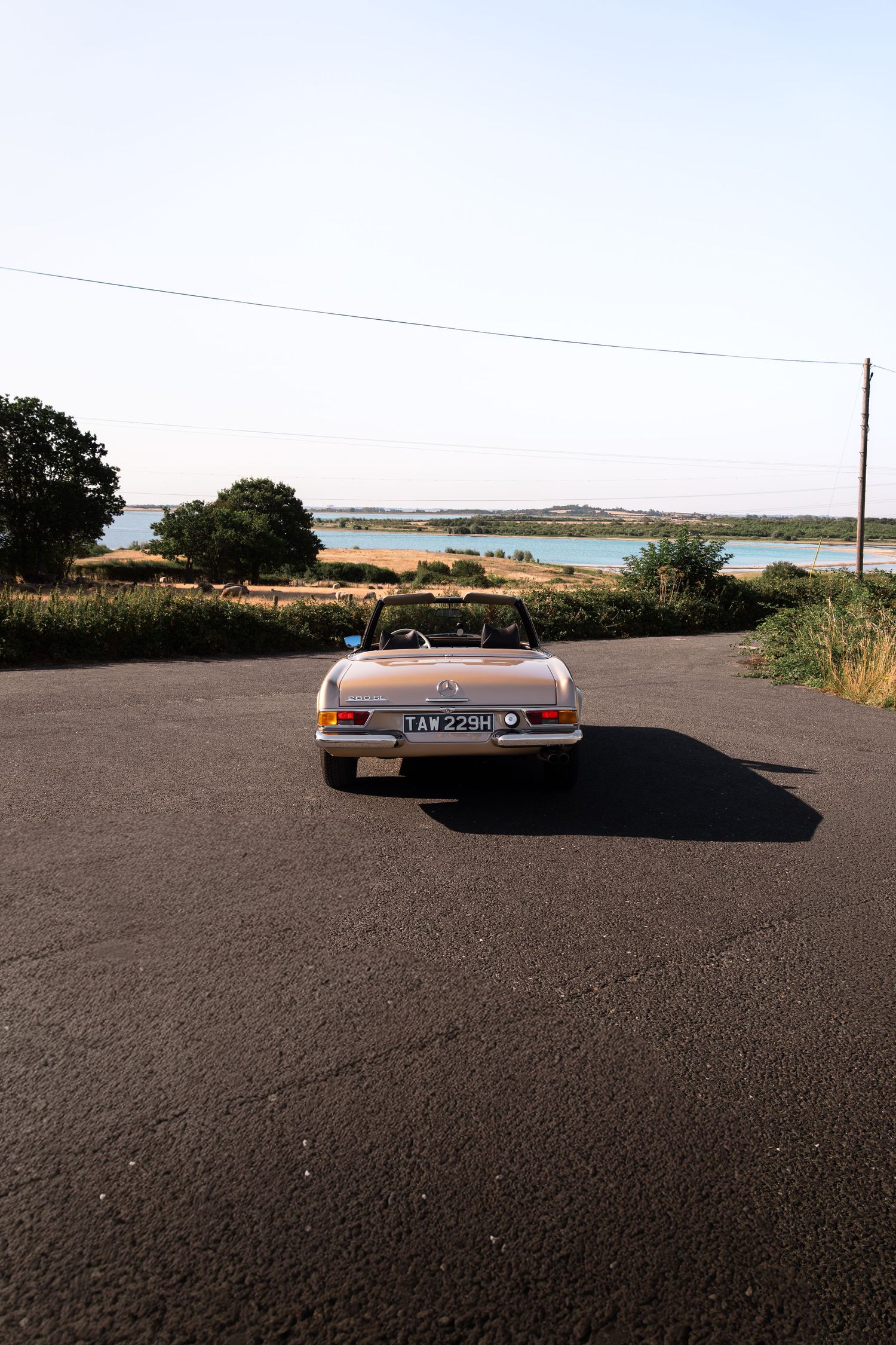 Vintage convertible car parked on a rural road with scenic lake and trees in background on a clear day.