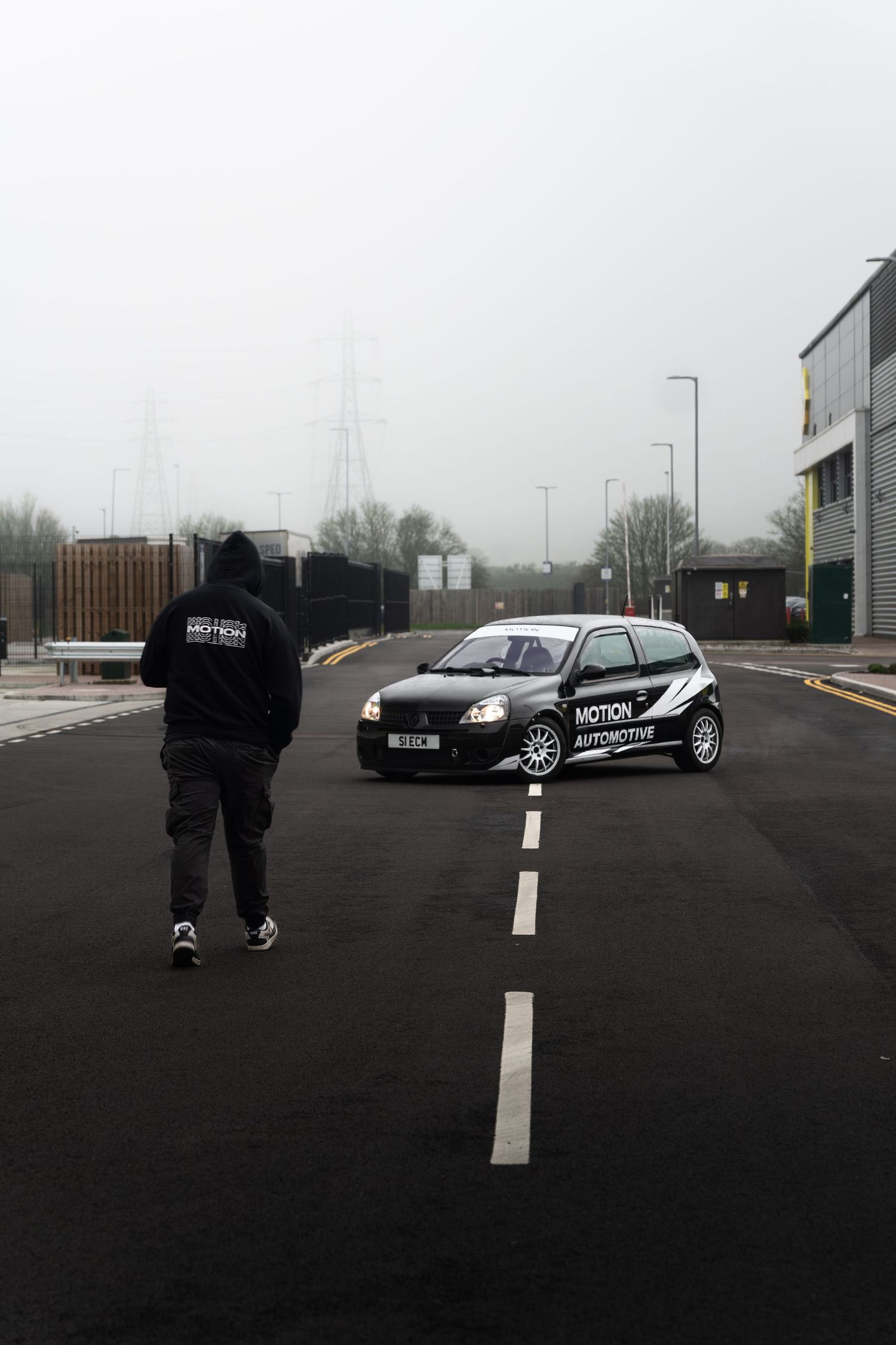 Person in Motion Automotive hoodie walking toward a black branded Renault Clio on a foggy industrial road.