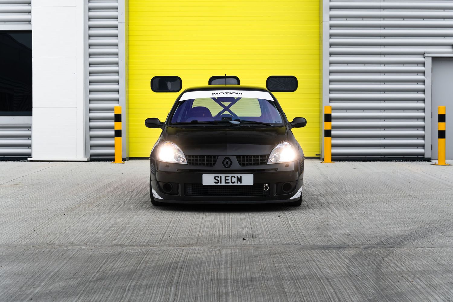 Front view of a black Renault Clio race car with S1ECM plate parked in front of yellow industrial garage doors.