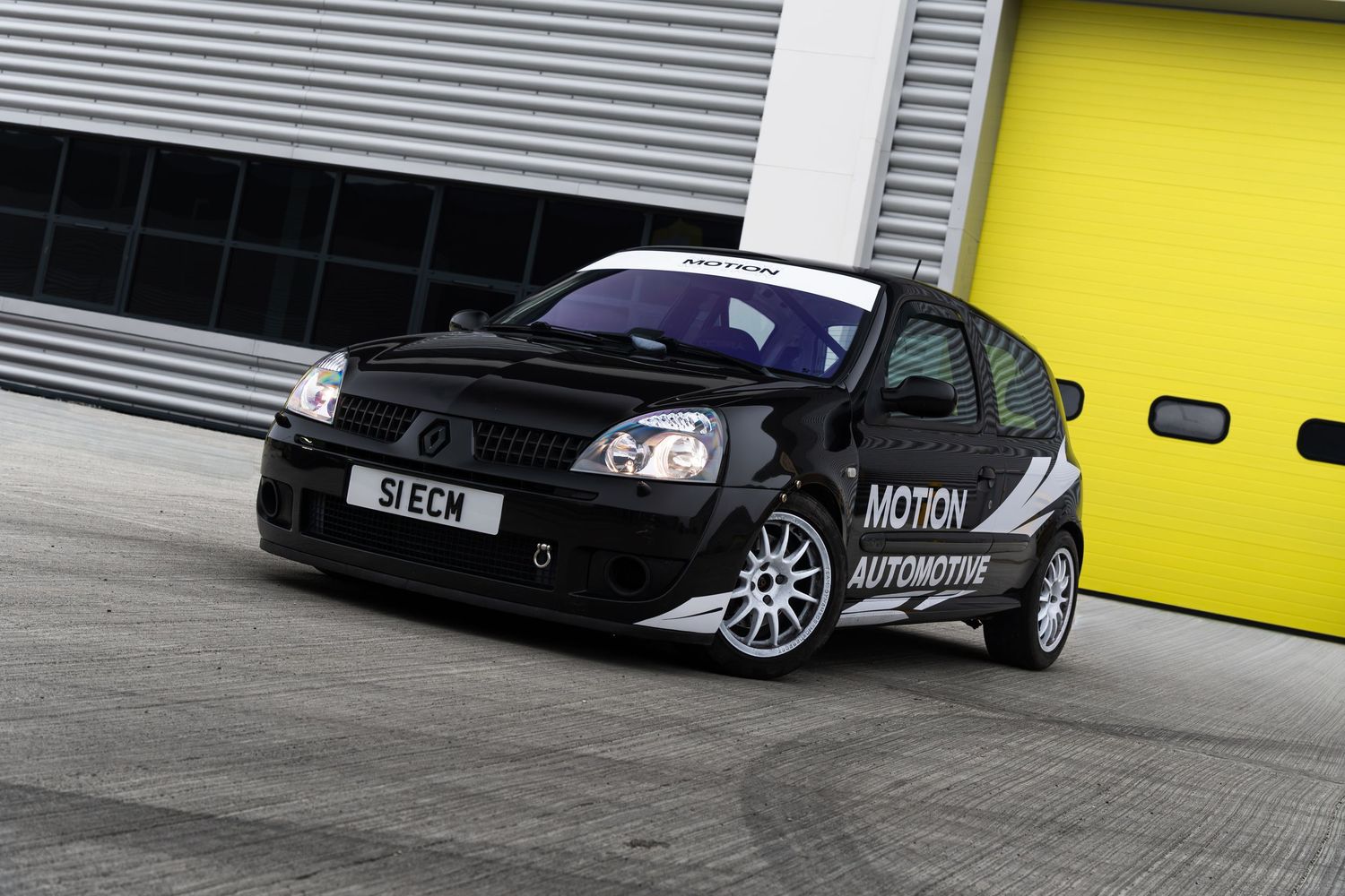 Black Motion Automotive racing Renault Clio with white decals parked outside yellow garage doors on a race track.