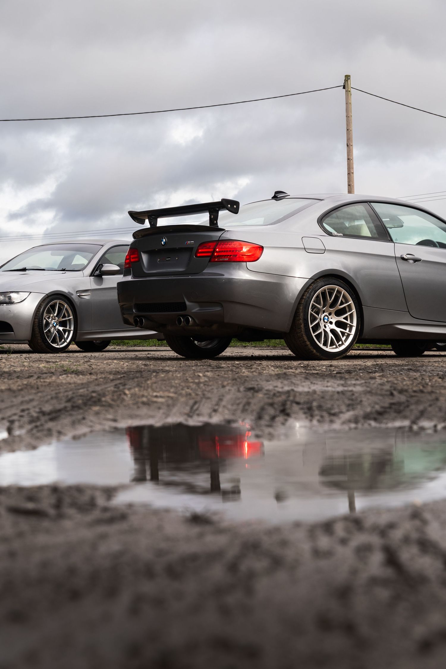 Rear view of a gray BMW M3 E92 with large GT wing reflected in a muddy puddle on overcast day.