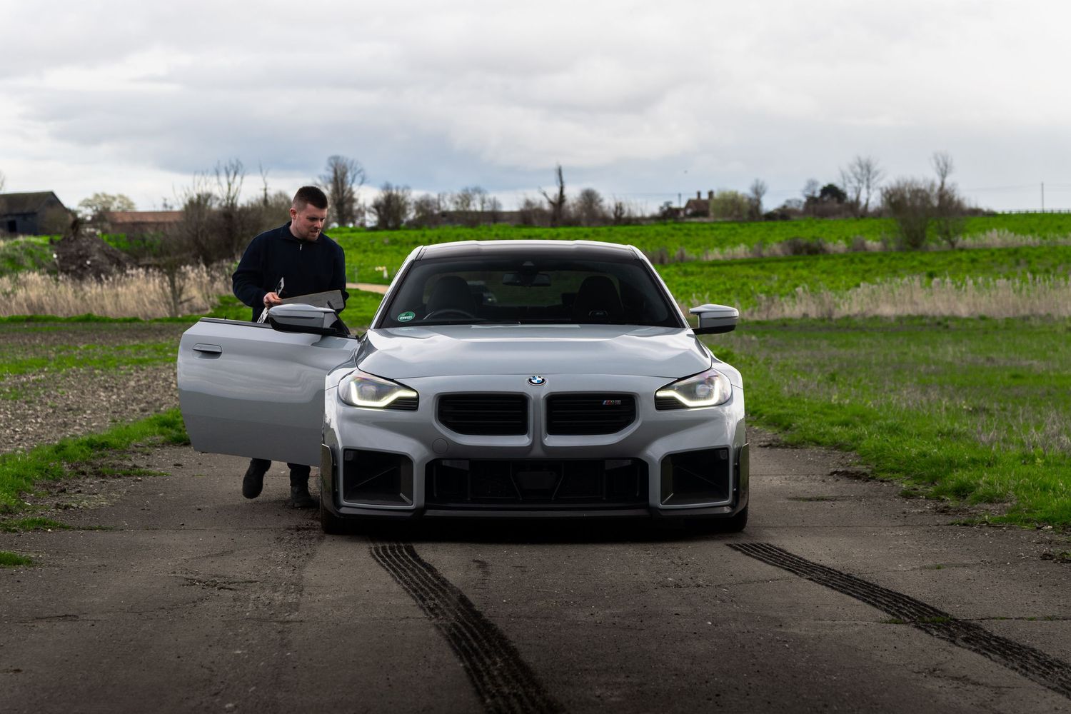 Man standing beside a white BMW M2 on a rural airfield with green fields.