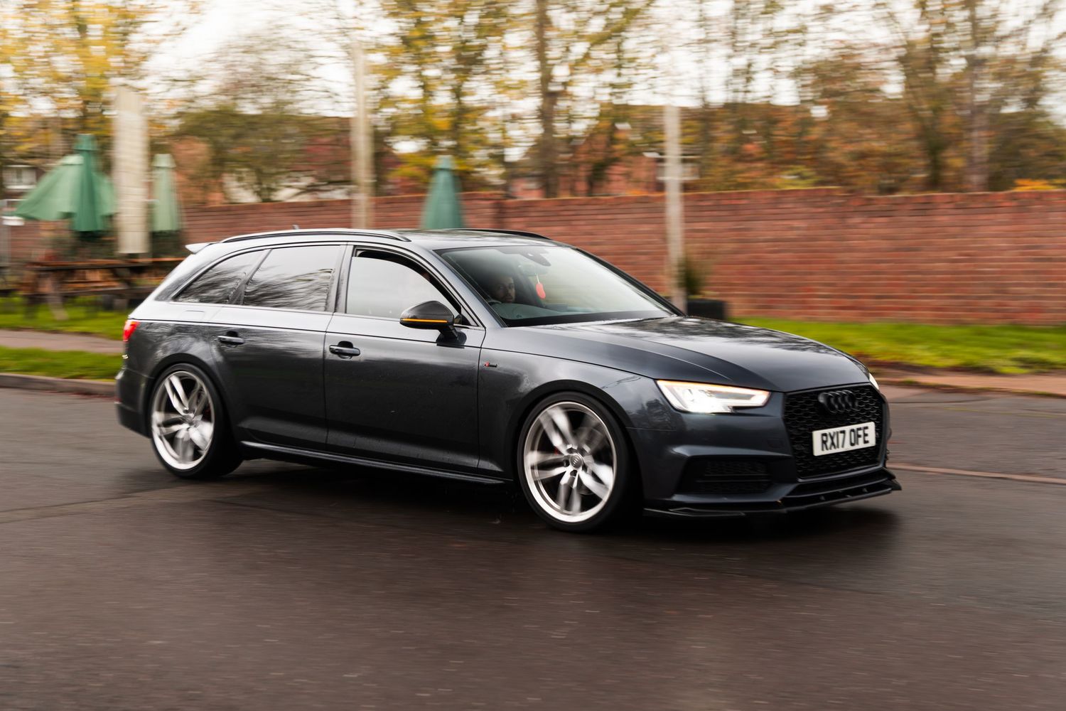Dark grey Audi A6 Avant estate car driving on wet UK road surrounded by autumn trees.