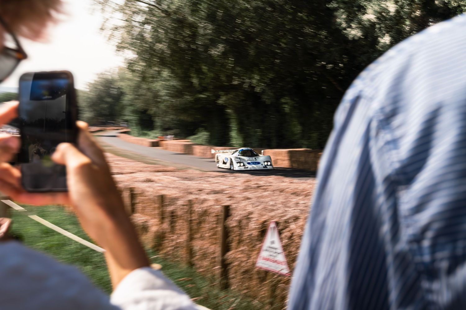 Spectator films a white racing car speeding along a hillclimb course lined with trees and dirt banks.