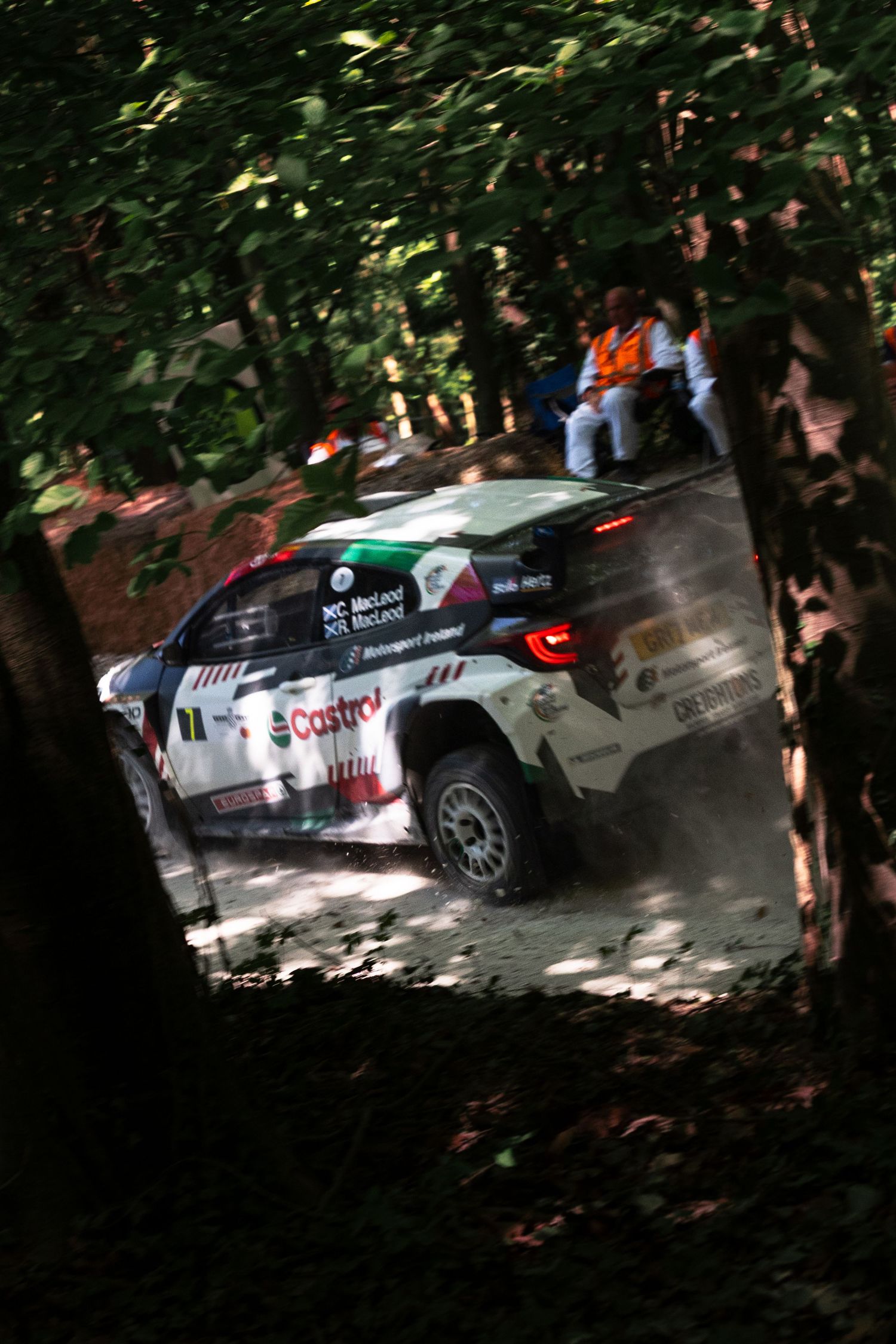 Rally car with Castrol sponsorship racing through a forest stage, kicking up dust on a dirt track.