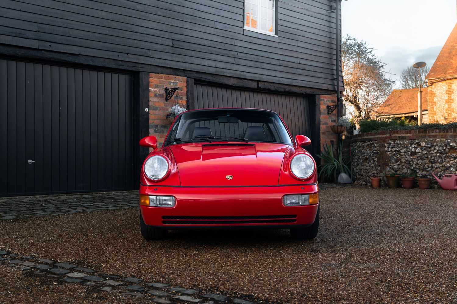 Straight-on front view of a red Porsche 911 Carrera 2 Targa parked on a gravel driveway at dusk.