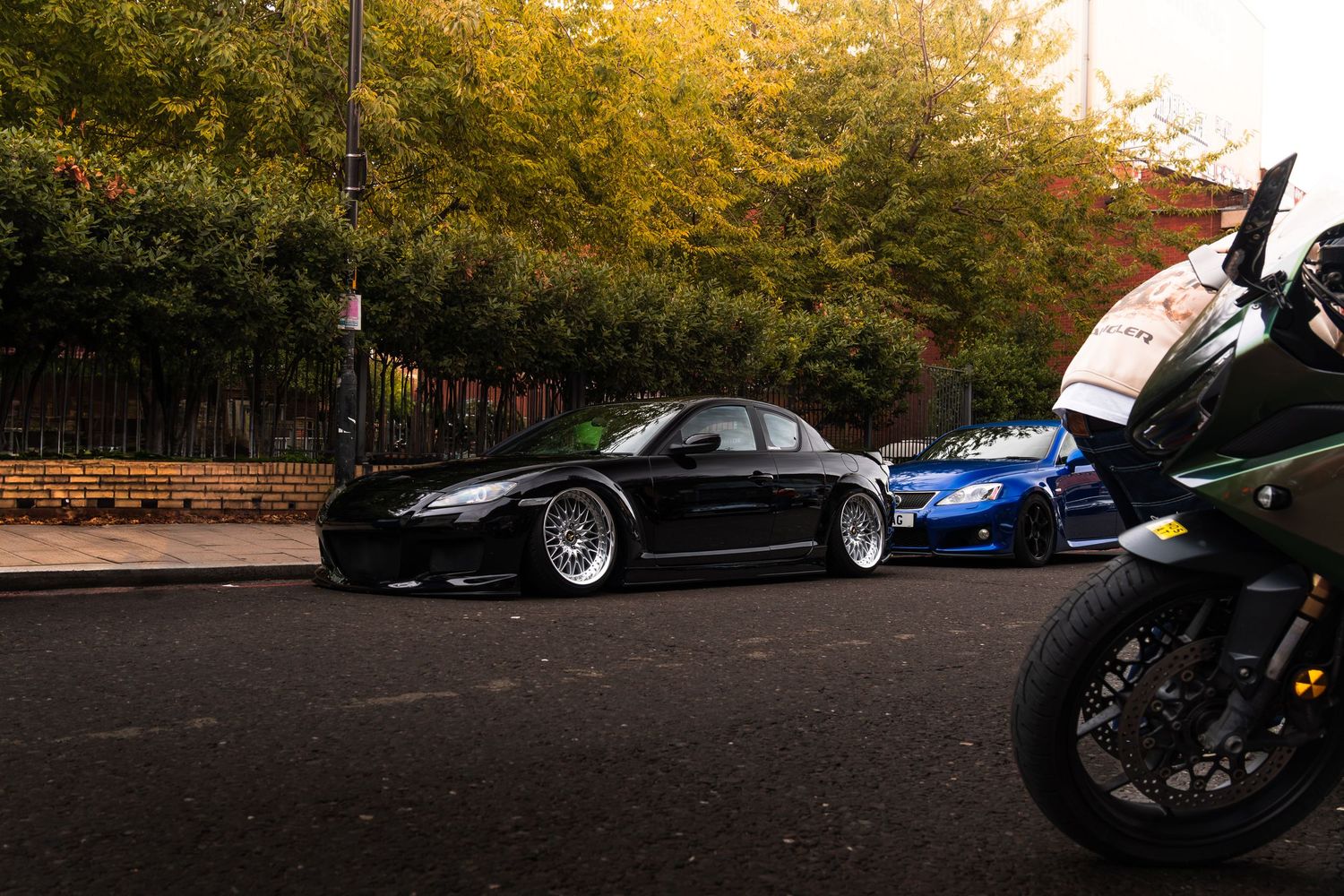 Two lowered black sports cars parked on a street beside a motorcycle during autumn.