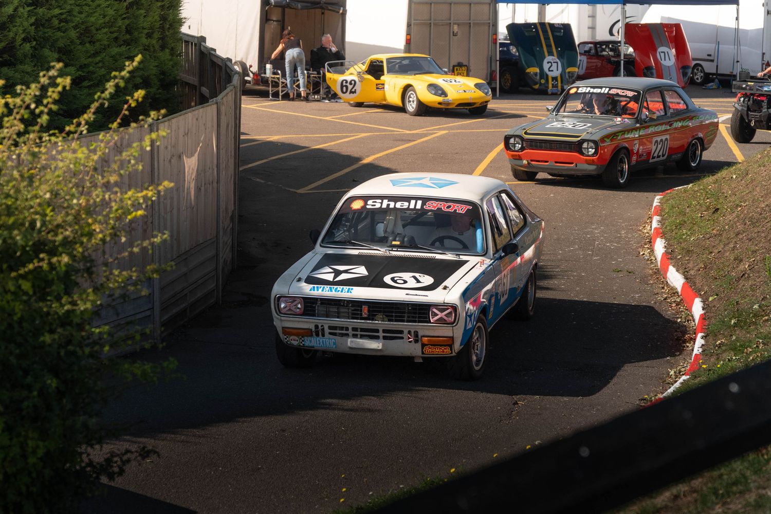 Classic Shell-sponsored racing car navigating paddock area with vintage cars following behind.