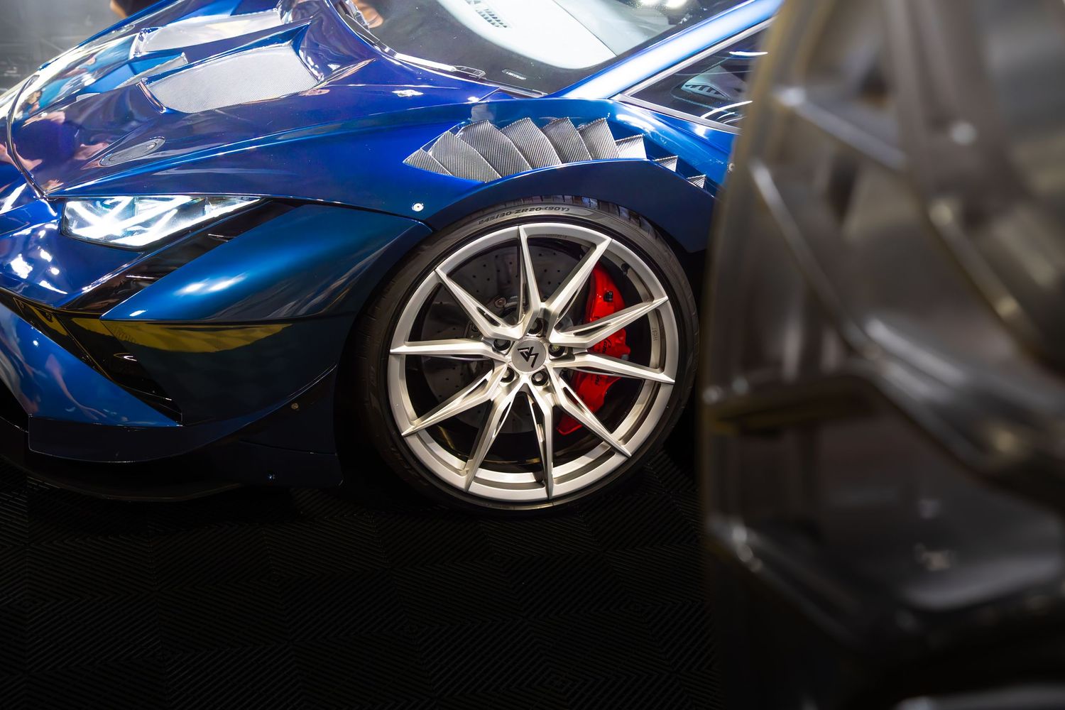 Close-up of a silver multi-spoke wheel with red brake caliper on a blue supercar.