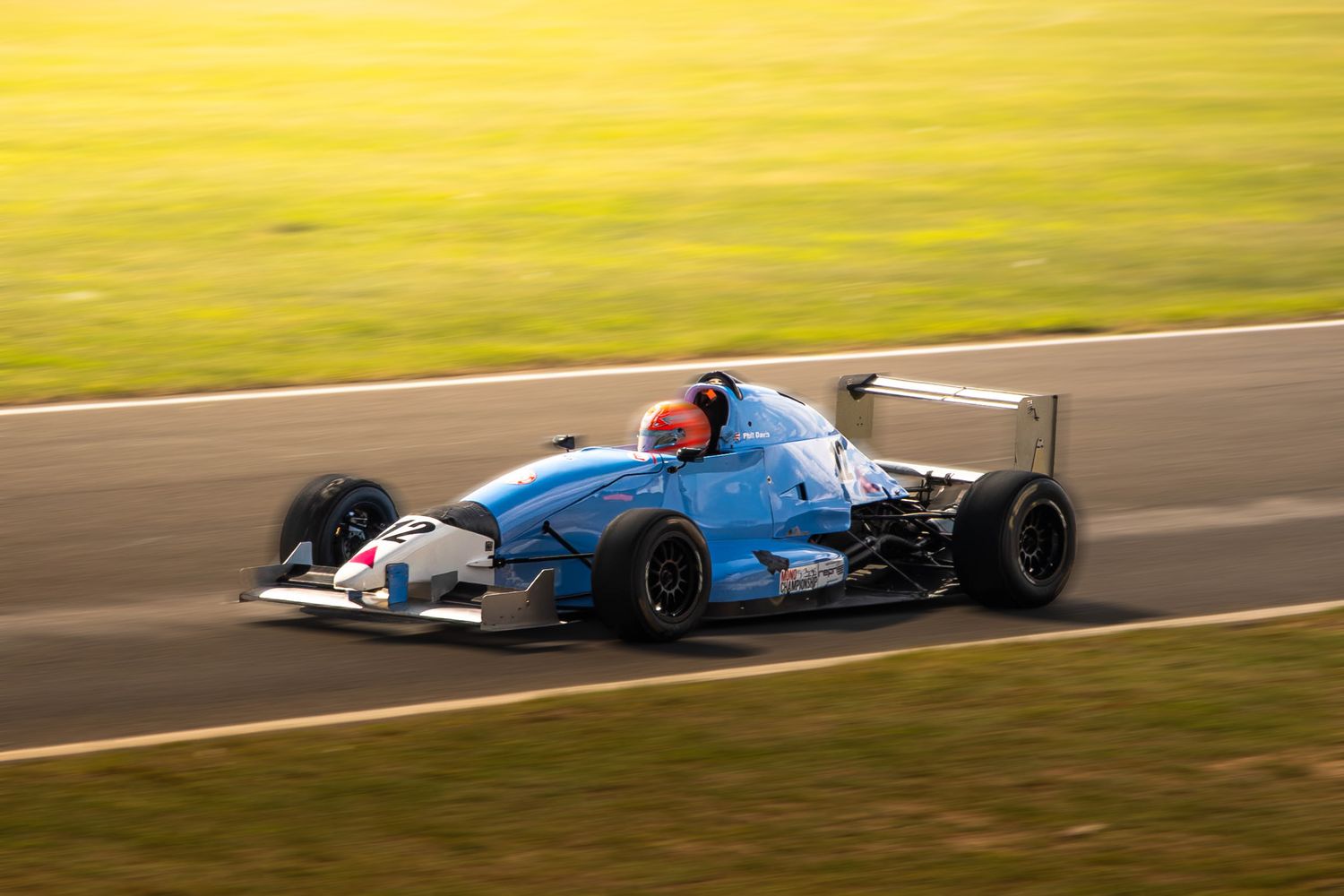Blue Formula racing car speeds along a sunlit race track during a motorsport event.