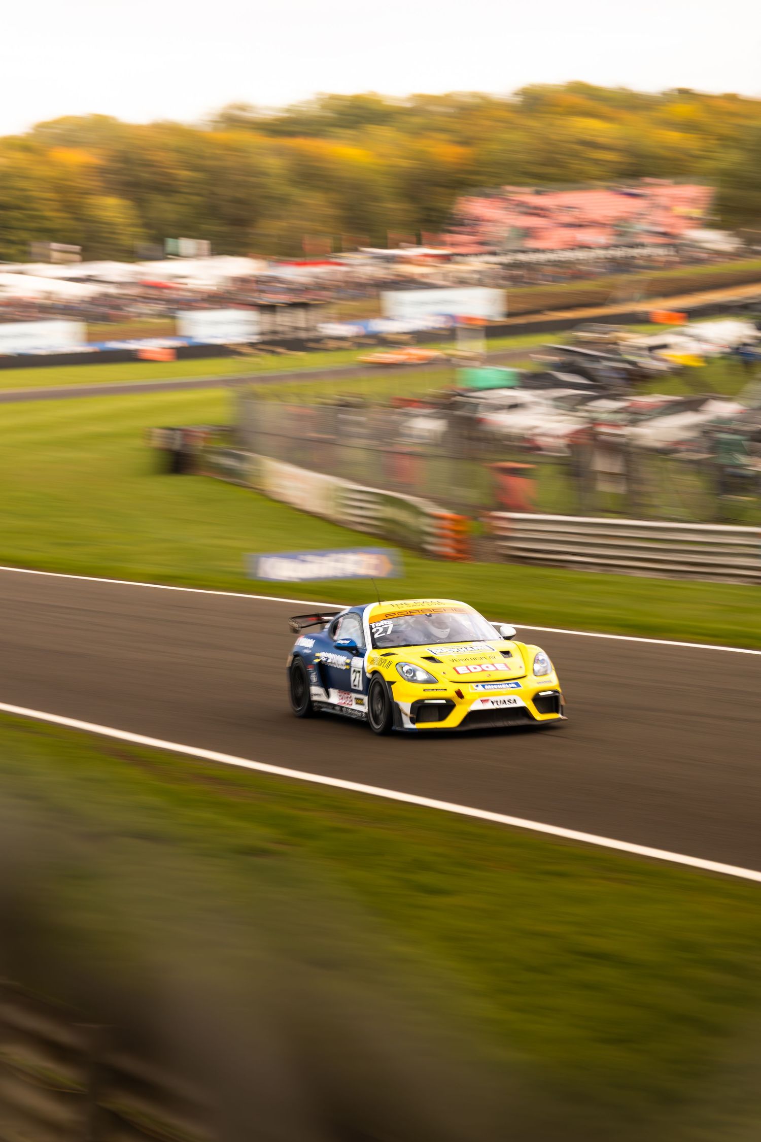 A yellow Porsche Cayman GT4 race car speeds along the track at a motorsport circuit.