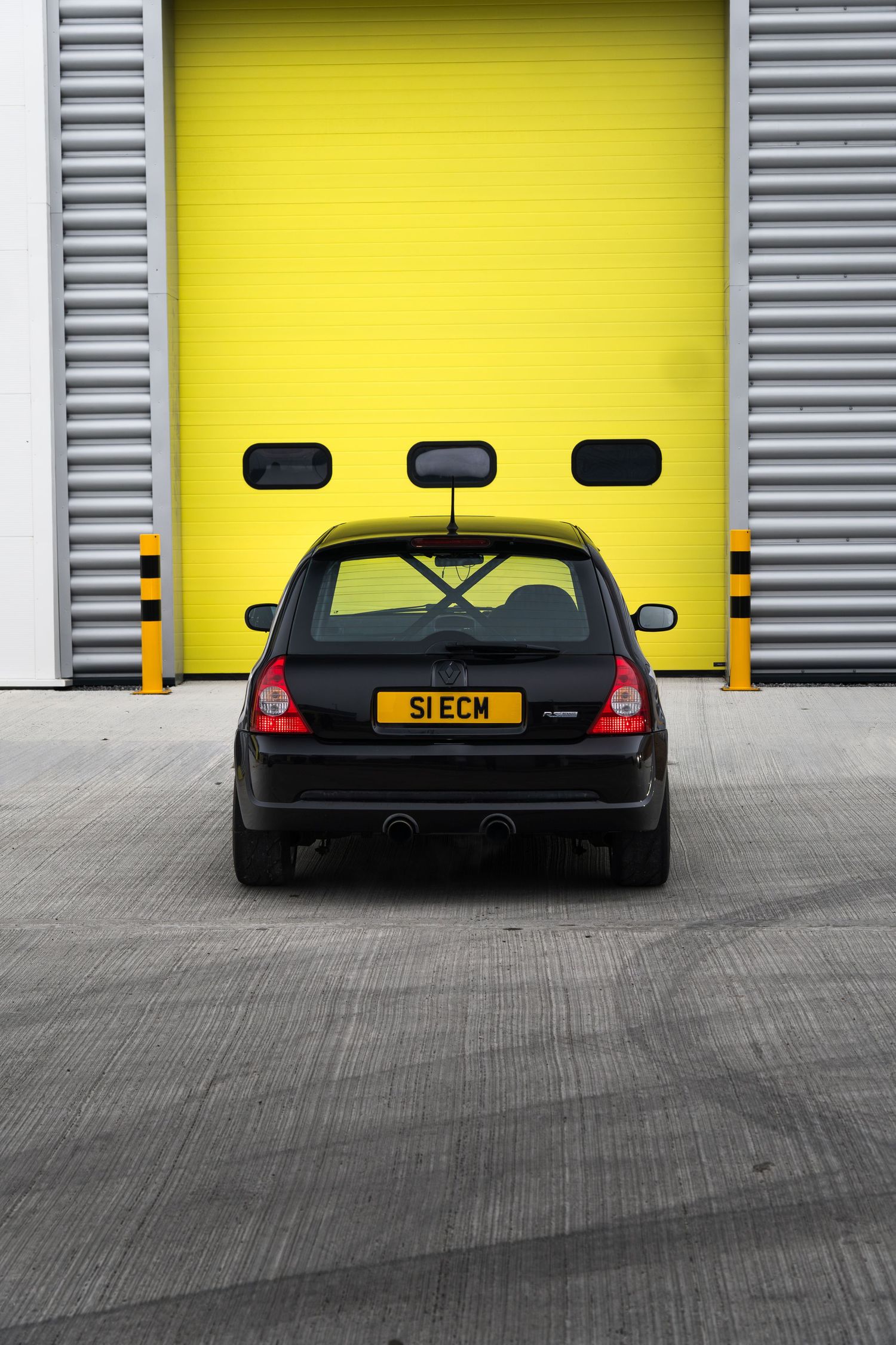 Black Renault Clio S1ECM parked centrally in front of a vivid yellow industrial garage roller door.