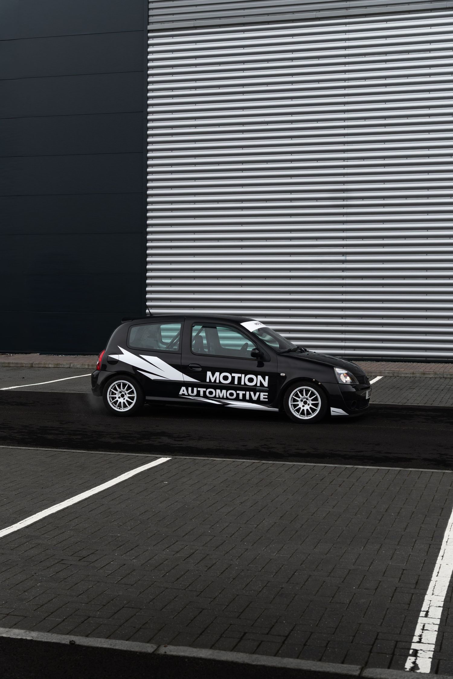 Black Motion Automotive Renault Clio parked on a brick-paved area beside a corrugated metal industrial wall.