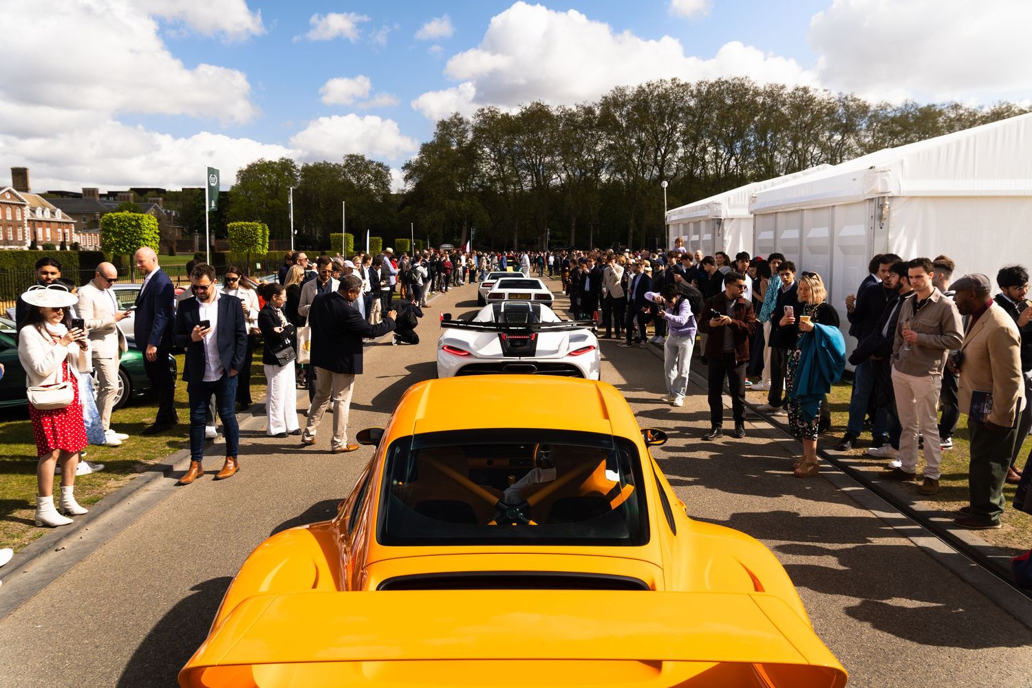Row of supercars including orange Porsche on display at outdoor automotive show with large crowd.