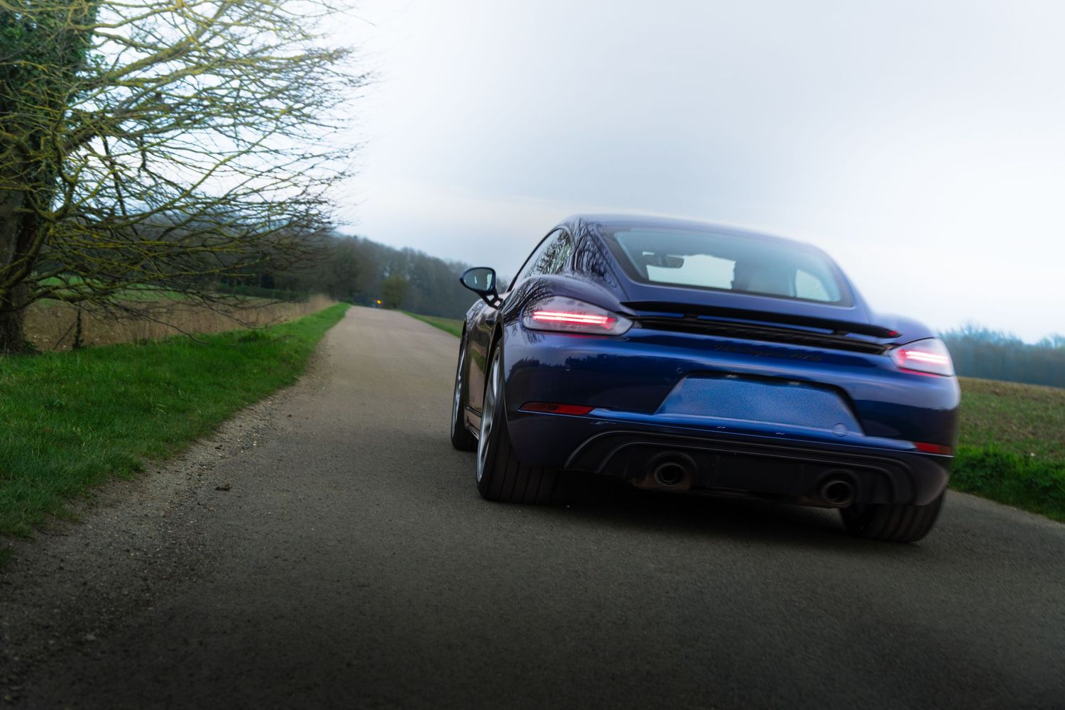 Rear view of blue Porsche 718 Cayman GTS on country road showing dual exhausts and diffuser.