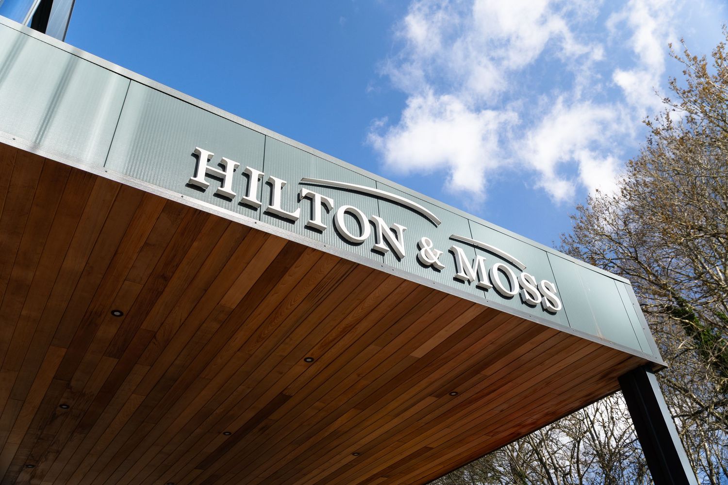 Hilton and Moss dealership entrance sign mounted on a wooden canopy structure against a blue cloudy sky.