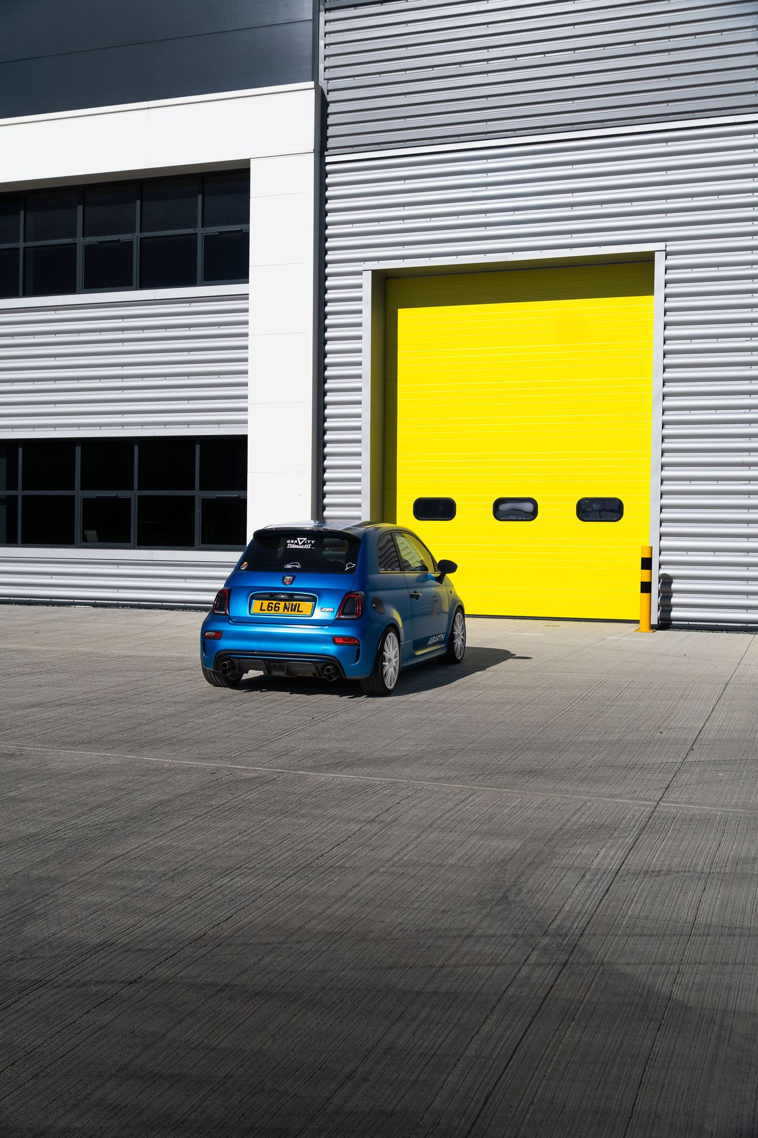 Blue Abarth 500 hatchback parked outside a grey industrial building with a yellow garage door.