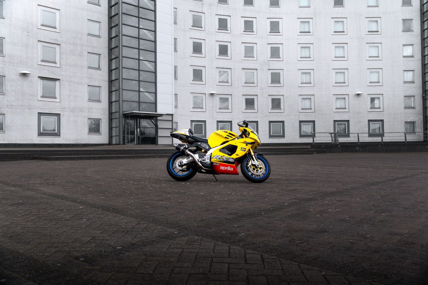 Yellow Aprilia RSV superbike parked at distance on dark cobblestones in front of a white building.